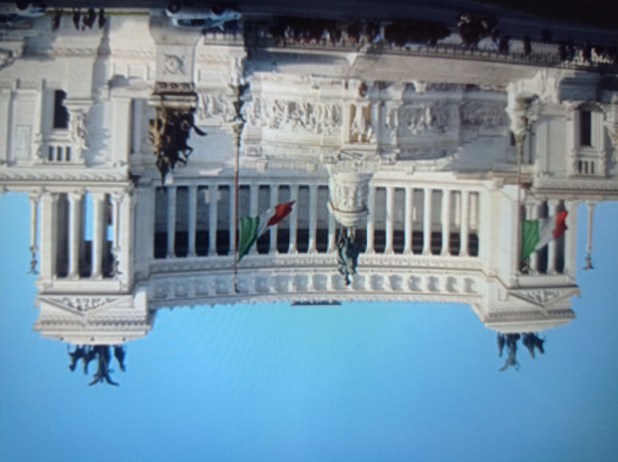 The image shows a large, white neoclassical monument against a clear blue sky. The monument features numerous columns, statues, and relief sculptures. Two Italian flags are prominently displayed on flagpoles in front of the building. In the foreground, a street is visible with several cars and a crowd of people gathered. The overall impression is that of a significant historical or civic landmark. However, there is no information in the image to suggest this location is in Mukachevo, Ukraine.