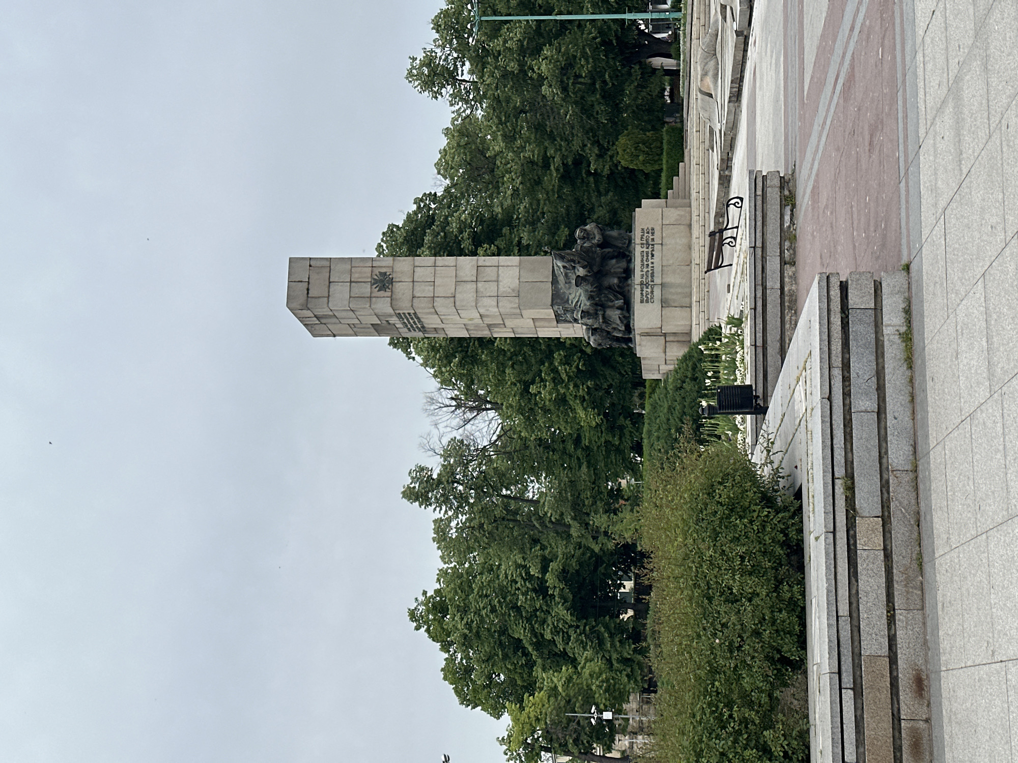 The image depicts the Monument of Glory located in the city of Vidin, Bulgaria. The monument consists of a tall, rectangular stone structure with a square base, adorned with a symbol. An artistic bronze sculpture of people with a flag is on one side of the monument. A marble inscription is displayed. The monument is surrounded by trees, bushes, a paved walkway, and low stone walls. The sky is overcast.