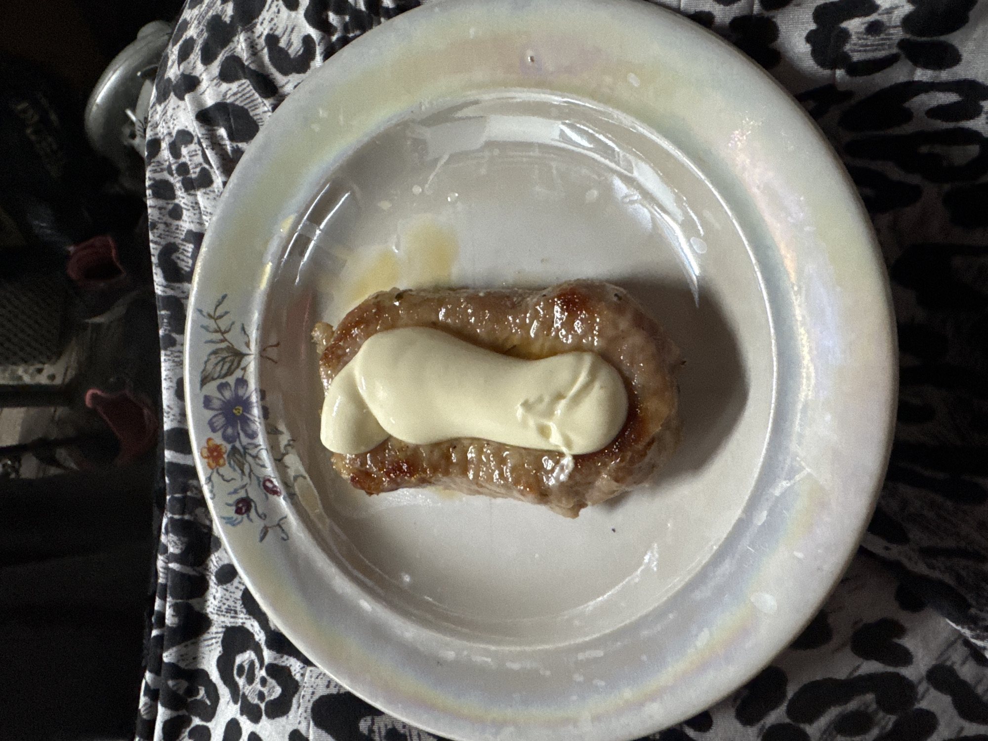 An overhead shot shows a single, cooked sausage on a white, iridescent plate. A dollop of pale yellow sauce, resembling mayonnaise, is squeezed on top of the sausage in a vertical line. The plate has a decorative floral rim. The plate rests on a black and white leopard print fabric. The background is dark and out of focus, with hints of red and gray objects. There is no identifiable location in the city of Chaikovsky, Russia.