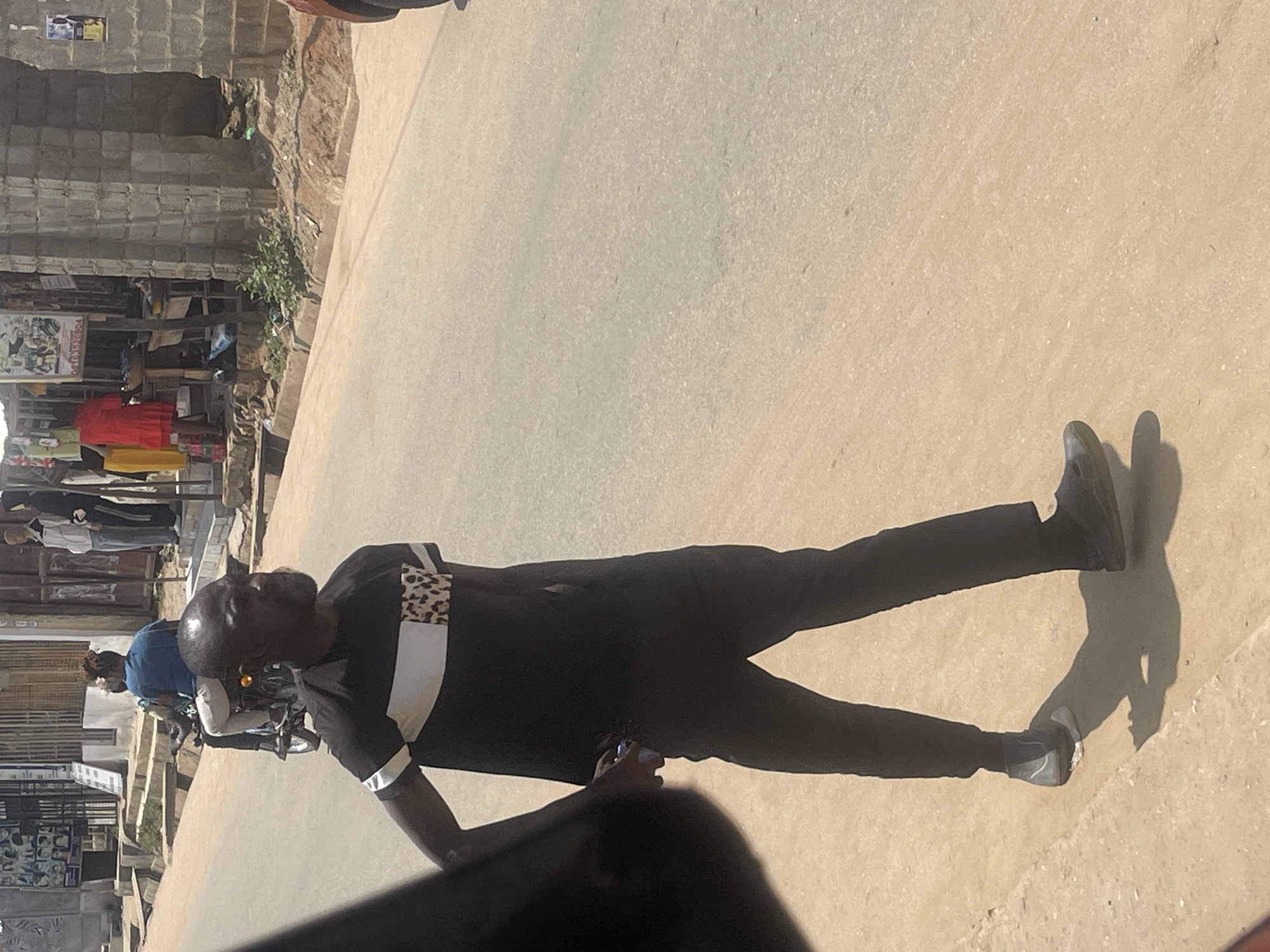 A man is walking on a dirt shoulder next to a paved road in Abuja, Nigeria. He is wearing a black t-shirt with white and leopard print accents, black pants, and dark shoes. He is holding a cell phone in his right hand. In the background, a motorcycle carrying two people is driving on the road. On the left side of the image, there are buildings and stalls that appear to be shops. The sky is bright, suggesting it is daytime.