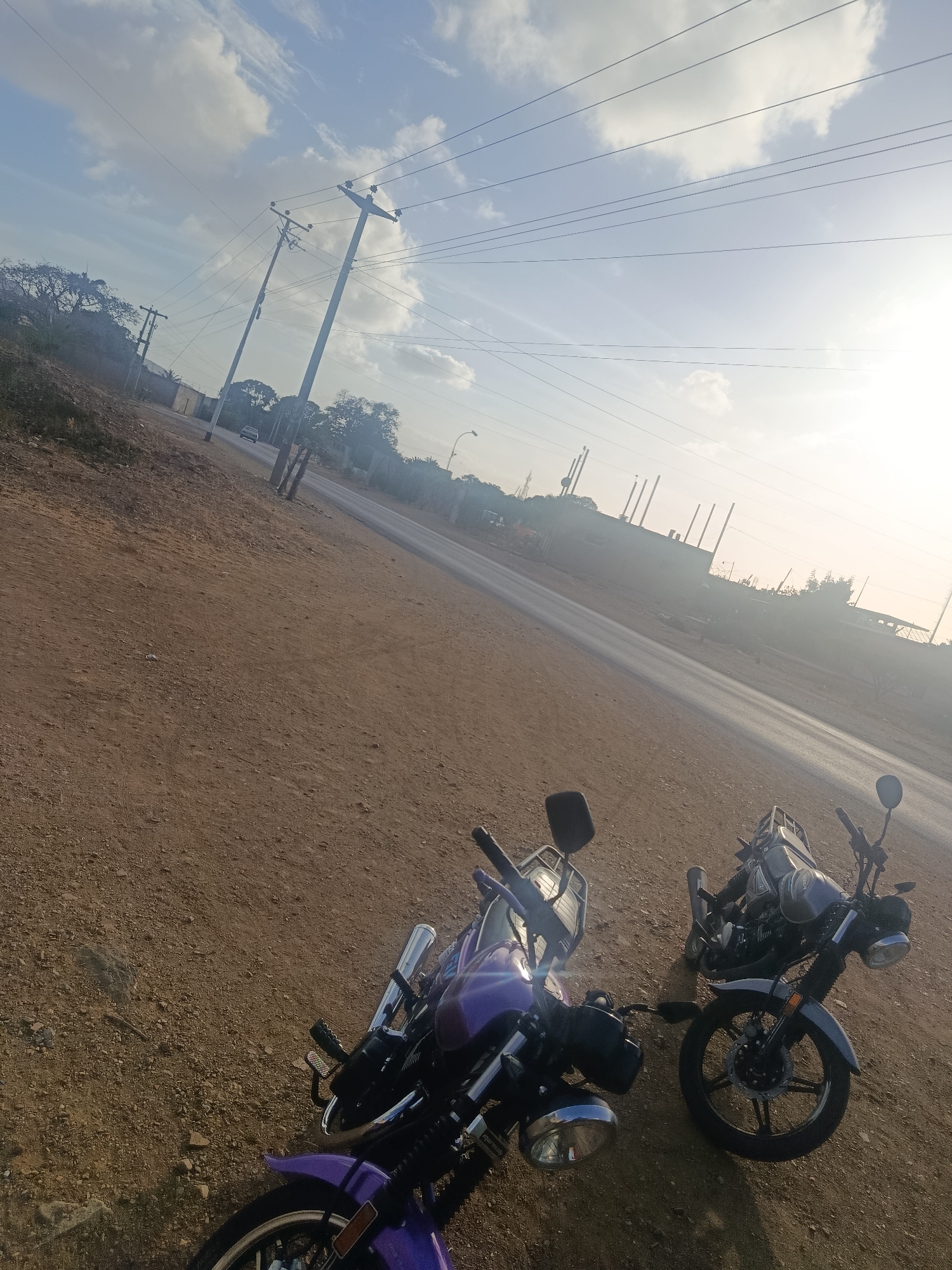 The image depicts a street scene in Juan Griego, Venezuela, featuring two motorcycles parked on a dirt shoulder adjacent to a paved road. The motorcycle on the left is purple, while the one on the right is gray. Overhead power lines stretch across the sky, supported by utility poles. In the background, a road leads to distant buildings. The weather appears sunny with some clouds in the sky. The time of day is either early morning or late afternoon, judging by the soft light and long shadows.