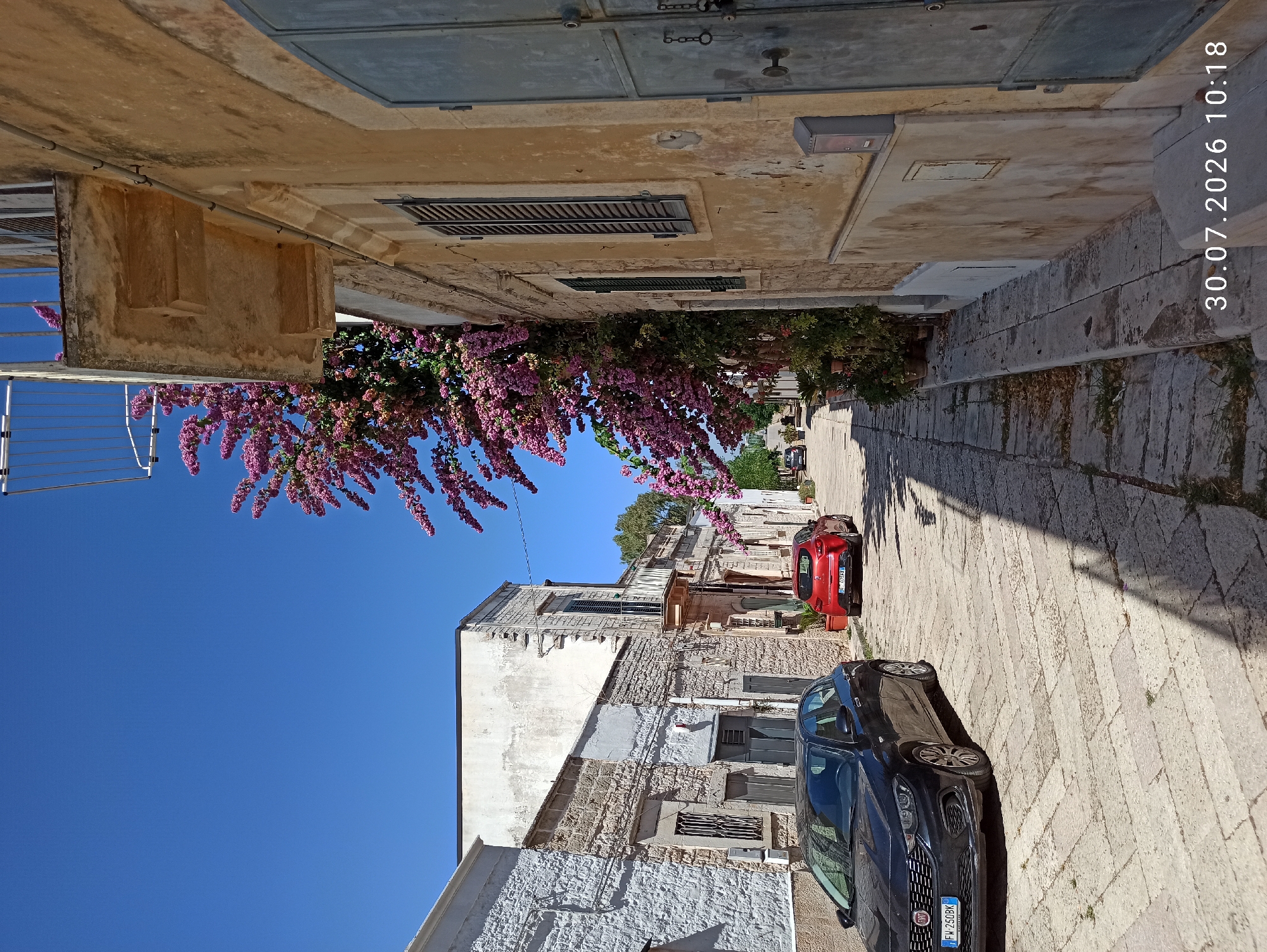 A narrow street in Terlizzi, Italy, is captured on a sunny day, with cobblestones paving the ground. A black Fiat, license plate FW-250BK, is parked on the left side of the street. A red Fiat is parked further down the street. Buildings line both sides of the street, some featuring stone walls and white plaster. A balcony with a clothesline extends from one of the buildings on the right. Vibrant pink bougainvillea cascades down a wall on the right side of the street, adding a splash of color to the scene. A blue metal door with chain detailing is visible on the building on the right. The sky is a clear, bright blue. The timestamp "30.07.2026 10:18" is overlaid on the bottom right corner of the image.