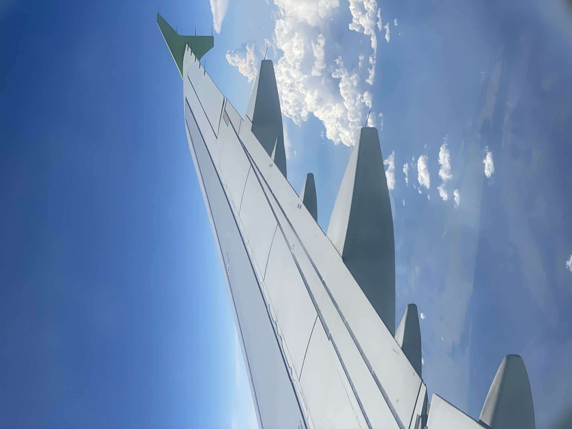 This media file is a photograph taken from an airplane window, looking upwards and outwards. The primary subjects are the wing of the airplane and the sky above. The airplane's wing dominates the lower portion of the image, appearing sleek and metallic with visible panel lines. The wing curves upwards and to the right, with several winglets extending upwards from its surface. The tail fin of the airplane, painted a light green, is visible at the top left.
The setting is the sky, characterized by a clear, bright blue expanse. Scattered white, fluffy clouds are visible against the blue, some appearing dense and voluminous while others are wispy. The sun is not directly visible, but its presence is indicated by the bright illumination and slight glare in the lower left part of the image, suggesting it is daytime.
Below the clouds, a hazy terrestrial landscape is visible, appearing somewhat abstract due to the altitude and possibly atmospheric conditions. This landscape suggests the airplane is in flight over land. There are faint lines and shapes on the ground, possibly fields or roads, but they are not clearly distinguishable.
The overall impression is one of travel and perspective from a significant altitude. The image conveys a sense of openness and the vastness of the sky. There are no people visible, only the man-made structure of the airplane's wing and the natural elements of the sky and land. No text is legible in the image. The context provided indicates the location as Сереброполь, Russia, although this specific location is not visually evident within the photograph itself.