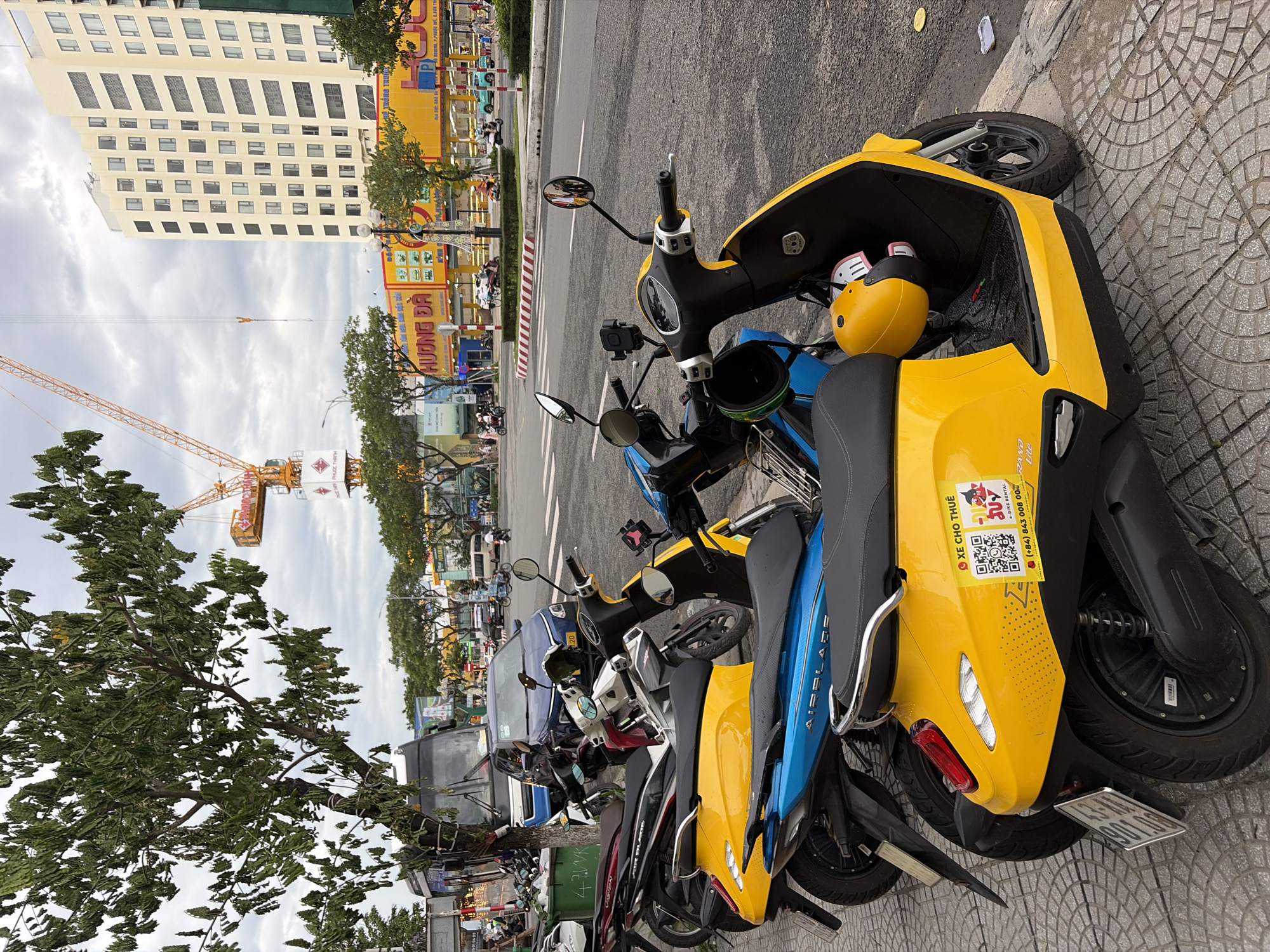 The image captures a street scene in Đà Nẵng, Vietnam, likely on a cloudy day. In the foreground, several parked motorbikes are visible. The closest motorbike is bright yellow, featuring a QR code sticker on its rear panel that reads "XE CHƠ THUÊ" (for rent) and "e-BIKE RENTAL," along with a phone number. A yellow helmet is stored in the front compartment of this yellow motorbike. To its left is a blue motorbike with "AIRBLADE" written on its side. Behind these, other motorbikes in various colors, including red and white, are parked.
The motorbikes are lined up on a sidewalk paved with grey interlocking bricks. Beyond the sidewalk is a street with traffic, including a blue car and a bus. In the middle ground, a large yellow building with many windows stands prominently. In front of this building are vibrant yellow and red signs, one of which appears to be a store. Further in the background, a tall construction crane is visible against the overcast sky, suggesting ongoing development. Trees line the street, and the overall impression is a busy urban environment.