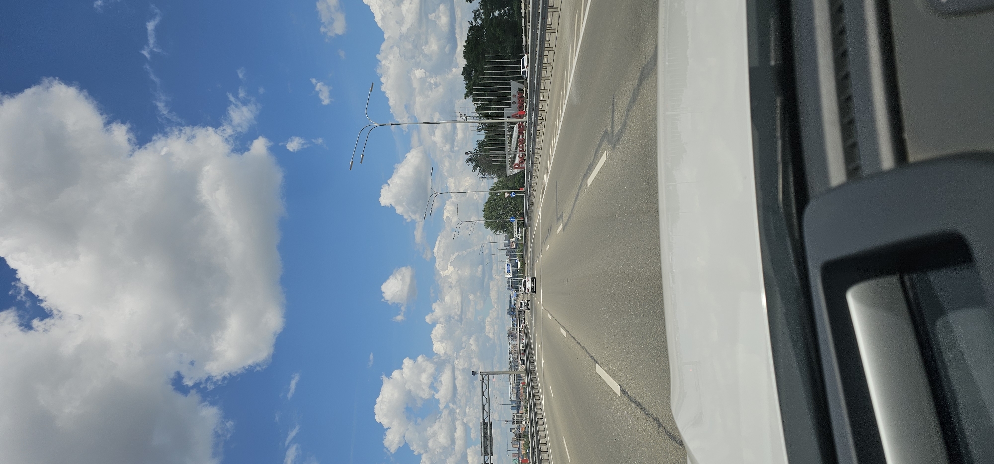 The image shows a view from inside a vehicle driving on a multi-lane highway under a bright blue sky with fluffy white clouds. The highway has clear lane markings and is lined with guardrails and streetlights. Several vehicles are visible on the road ahead, including cars and what appears to be a truck. On the right side of the highway, there is a large sign with Cyrillic lettering that reads "РОСНЕФТЬ" and appears to be an advertisement or landmark. Beyond the sign, there is a line of trees and some buildings in the distance. The foreground of the image shows the interior of the vehicle, including part of the dashboard. The scene appears to be captured during daylight hours. The specific location is unknown.