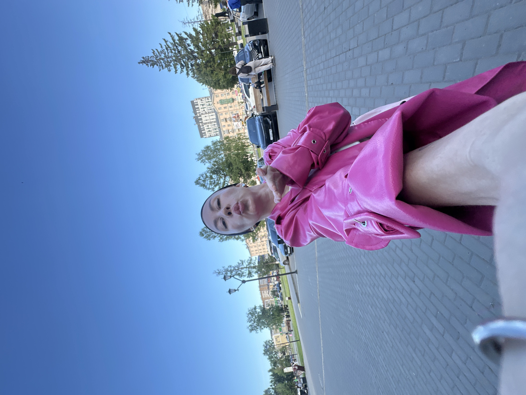 A selfie-style photograph, taken outdoors in Izhevsk, Russia, features a person in a bright fuchsia-pink, shiny long coat, posing with pursed lips for a "kissing" face. The person also wears a dark head covering, possibly a bandana or scarf. The image is taken from a low angle, looking slightly upwards. On the far right, a blurry arm or leg is visible in the foreground, suggesting the camera is held close.
The background reveals a clear, vibrant blue sky dominating the upper portion of the frame. Below the sky, an urban setting unfolds. A wide paved sidewalk or pedestrian area, made of light-colored rectangular bricks, stretches into the distance. Along the street, several cars are parked or driving, mostly dark-colored sedans. Green deciduous and coniferous trees are scattered along the pavement, providing shade. In the mid-ground, multi-story buildings with an older, European architectural style line the street. Streetlights are also visible among the trees.
The scene is brightly lit by natural sunlight, indicating a clear, sunny day, likely morning or afternoon. There is no visible text in the image. The overall mood is casual and playful, captured in a busy city environment.