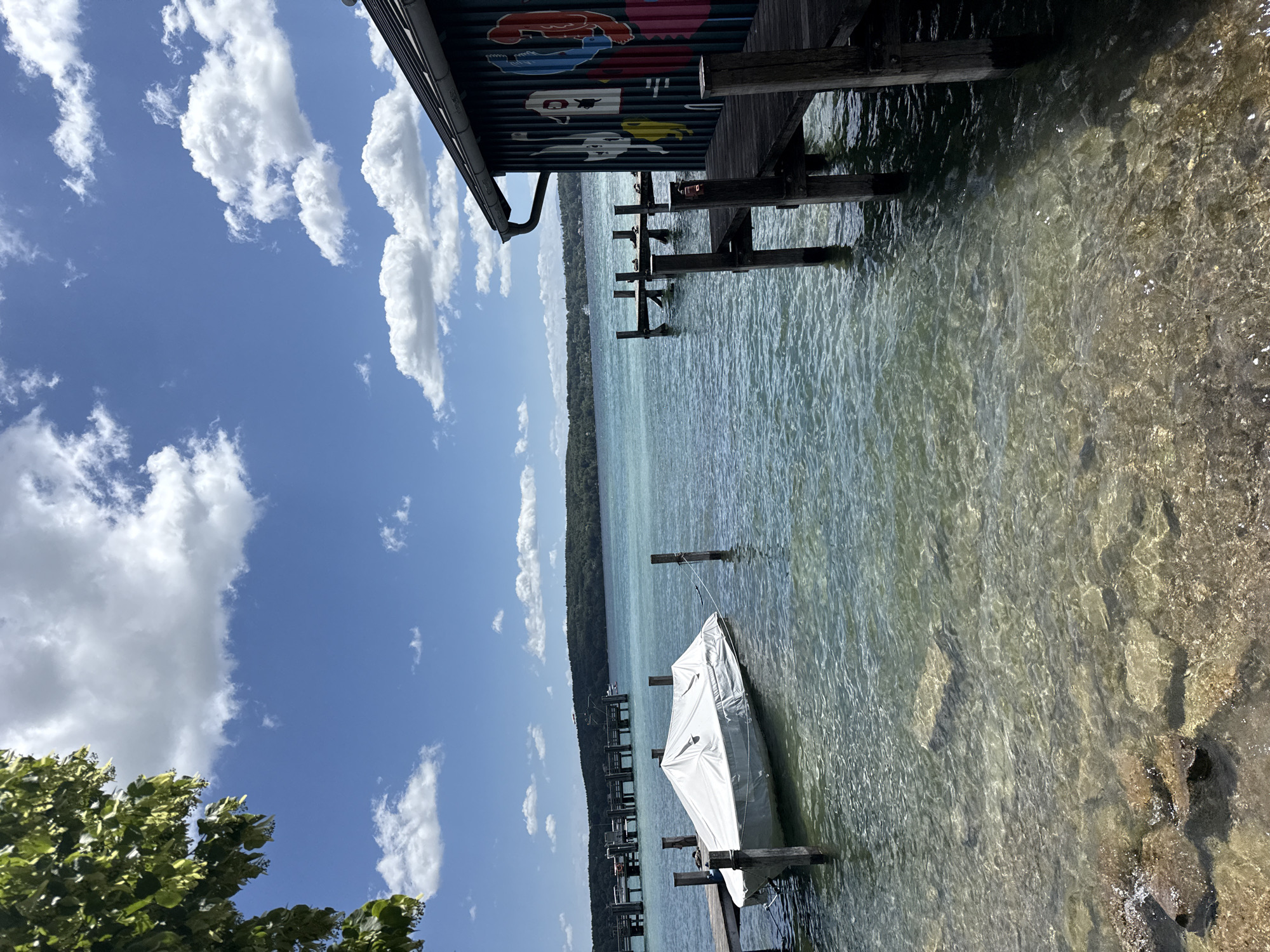 A bright, sunny day is captured in this image, likely taken in Starnberg, Germany. The scene depicts a clear, turquoise lake with a wooden pier in the foreground, leading to a boathouse adorned with colorful cartoonish figures on its side. A boat covered with a white tarp is docked by a smaller pier to the left, its reflection shimmering on the water's surface. The lake extends towards a lush, tree-covered shoreline in the distance, under a sky dotted with fluffy white clouds. The water is so transparent that the rocky lakebed is visible beneath its surface.