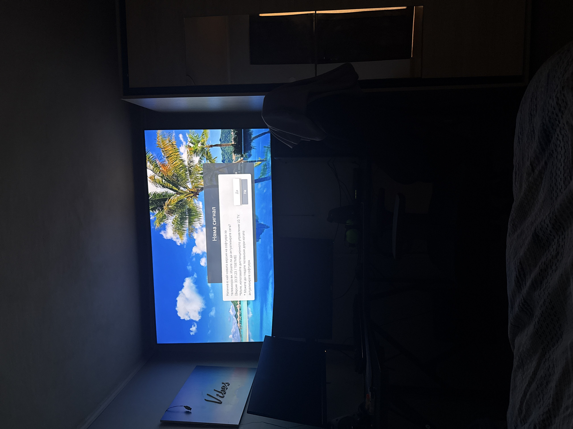 The image shows a television displaying a screen in Bulgarian that reads "No signal" at the top, with a message about a software update for the LG TV. The TV is showing a tropical beach scene in the background. In the bottom left of the frame, there's a partially visible sign that says "Vibes". The room is dark, with various objects, including what appears to be another monitor and possibly some audio equipment. A patterned blanket or fabric is visible on the right side of the frame. Due to the lack of landmarks, it is not possible to identify the location in Shumen, Bulgaria.