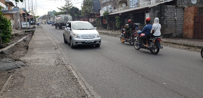 A silver sedan drives down a paved road in Keshero, Democratic Republic of the Congo. The road is in fair condition, with some cracks and potholes. The sidewalk to the left of the road is uneven and dirt, while the sidewalk on the right is paved and in good condition. A small truck is parked in the background on the left side of the road, and several people are visible, some on foot and others driving motorcycles. The scene is typical of a city in the developing world, with a mix of old and new vehicles and some signs of neglect in the infrastructure. There is a brick building and a few trees in the background on the right side of the image. The driver of the silver sedan is focused on the road. Three people are riding a red and black motorcycle toward the left of the image, the passenger on the back is wearing a white hooded sweatshirt.