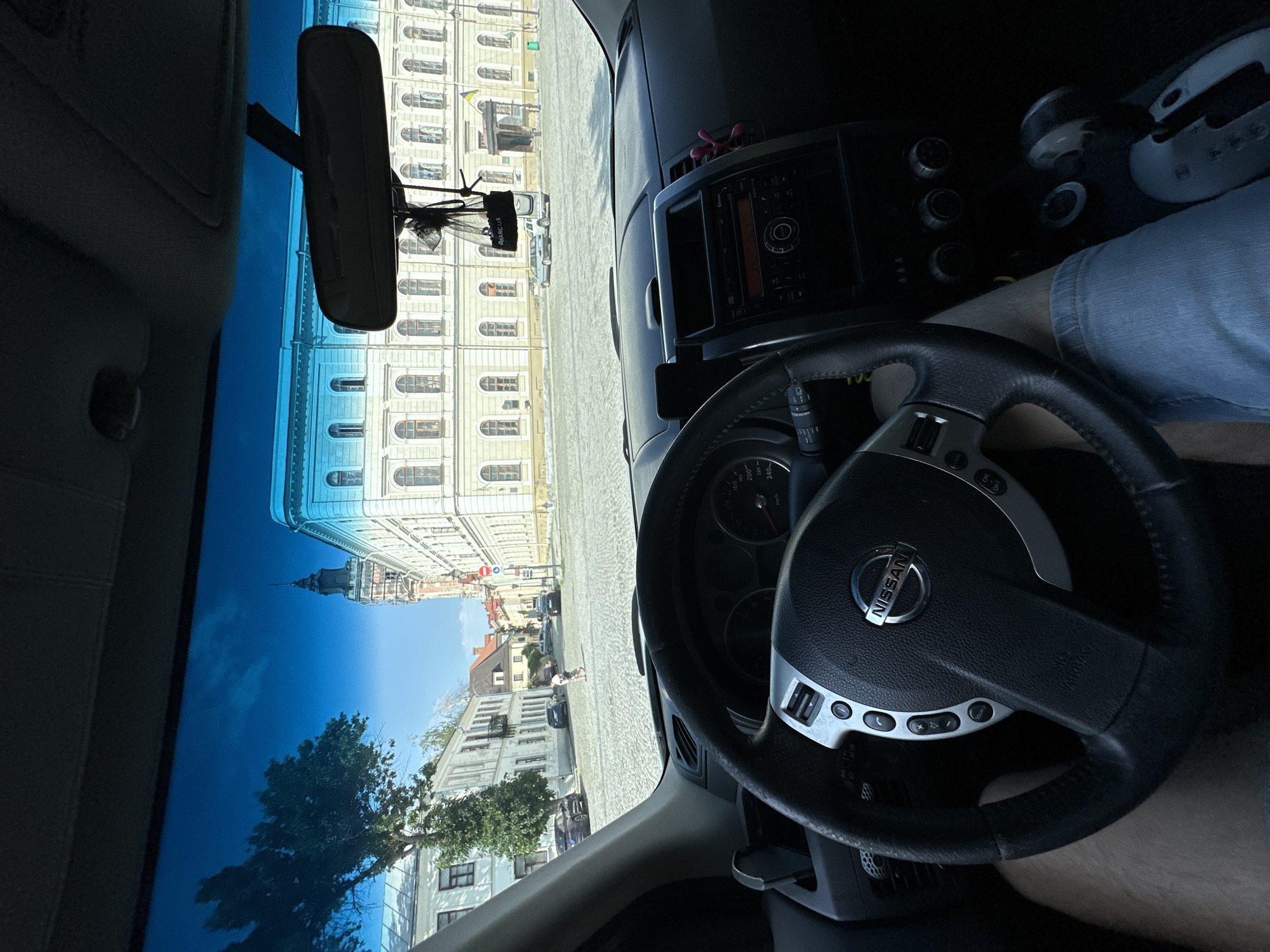 The image is a first-person view from inside a Nissan car, looking out through the windshield. The car is parked on a cobblestone square in front of an ornate, light-colored building with arched windows. A tall, dark-colored tower with a spire is visible to the left of the building. Several other buildings line the square, and trees are present. A few cars and a person are visible in the distance, walking across the square. The steering wheel of the car is prominent in the foreground, as is the dashboard with the radio and climate controls. A rearview mirror hangs from the windshield. A small dark-colored air freshener is hanging from the mirror. The sky is blue with scattered clouds. The scene appears to be set in the city of Kamianets-Podilskyi, Ukraine, likely near the Old Town or the castle. The overall lighting suggests it is daytime, possibly late morning or afternoon.