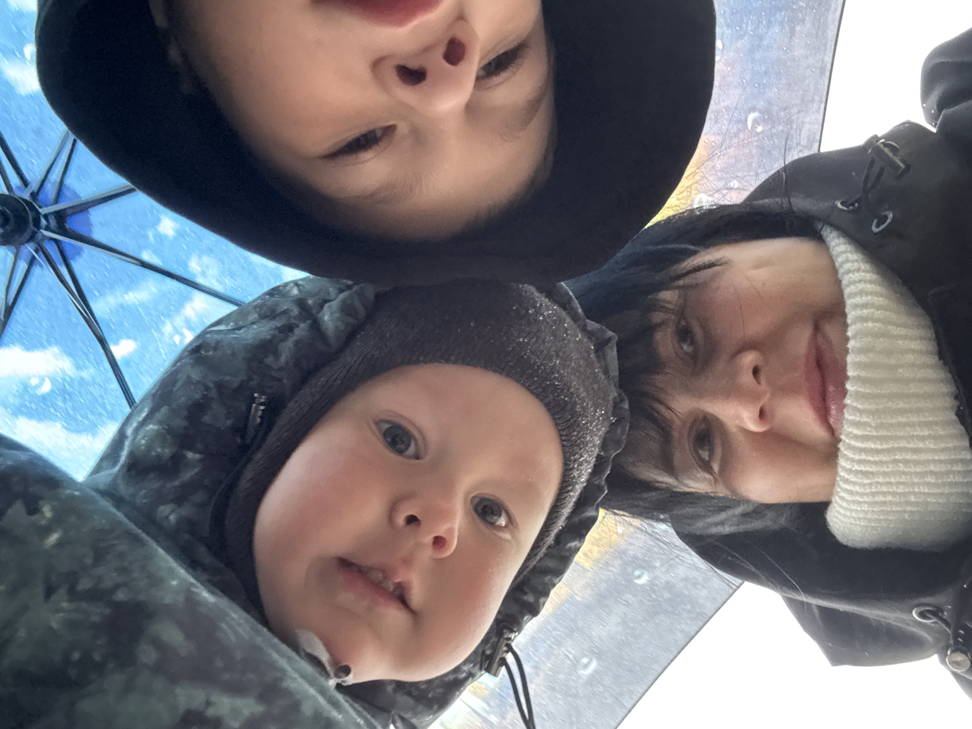 A selfie taken from a low angle shows three people and part of an umbrella. The umbrella's frame and translucent canopy, speckled with what appears to be raindrops, are visible. At the top of the frame, a person wears a dark hooded garment. Below and to the left is a young child wearing a dark, textured hat with a hood attached to a jacket. To the right is an adult with dark hair wearing a black jacket over a white sweater. The faces of all three people are prominent and close to the camera, implying they are huddled together, likely under the umbrella. The exact location in Zav'yalovo, Russia, is not discernible from the image.