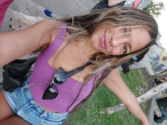 The image depicts a female individual taking a selfie in an urban park environment within Buenos Aires, Argentina, characterized by a large concrete wall featuring geometric graffiti. The primary subject, in the foreground, faces the camera with a direct gaze and a closed-mouth smile. She has long, wavy, dark hair with blonde highlights and light-brown skin. Facial features include a silver stud piercing on her right nostril and a silver labret piercing below her lower lip. She wears a purple sleeveless tank top and light blue denim shorts. Black sunglasses with round lenses are hanging by a silver chain around her neck, resting on her chest. A black handbag is visible on her right side. Her right arm is extended upwards and to the right, partially out of frame.
The background shows a public park scene. On the left, a concrete surface displays a blue plastic bottle and a light-colored bag. On the right, a grassy area contains a gnarled wooden pole. Several blurred individuals are visible in the mid-ground, including two seated on a concrete barrier or bench. The background is dominated by a tall concrete wall adorned with abstract geometric graffiti in green, purple, white, grey, and black. Trees and foliage are visible above the wall and in the distant background. The scene is illuminated by natural light.