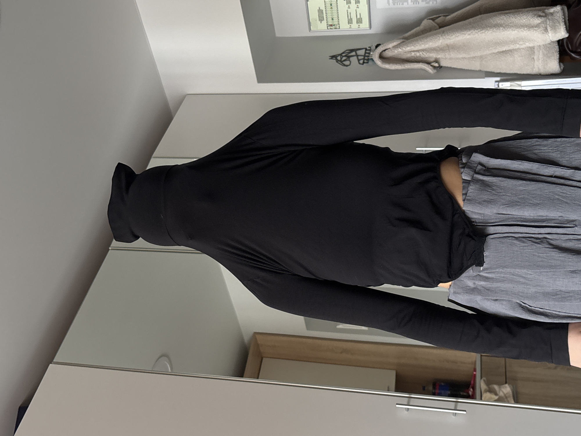 A person wearing a black turtleneck shirt and grey pants is standing in front of a mirrored wardrobe. The location is not identifiable as Perm, Russia. The wardrobe has white doors with silver handles. A building map is affixed to the wall above the wardrobe, along with a cream-colored coat hanging.