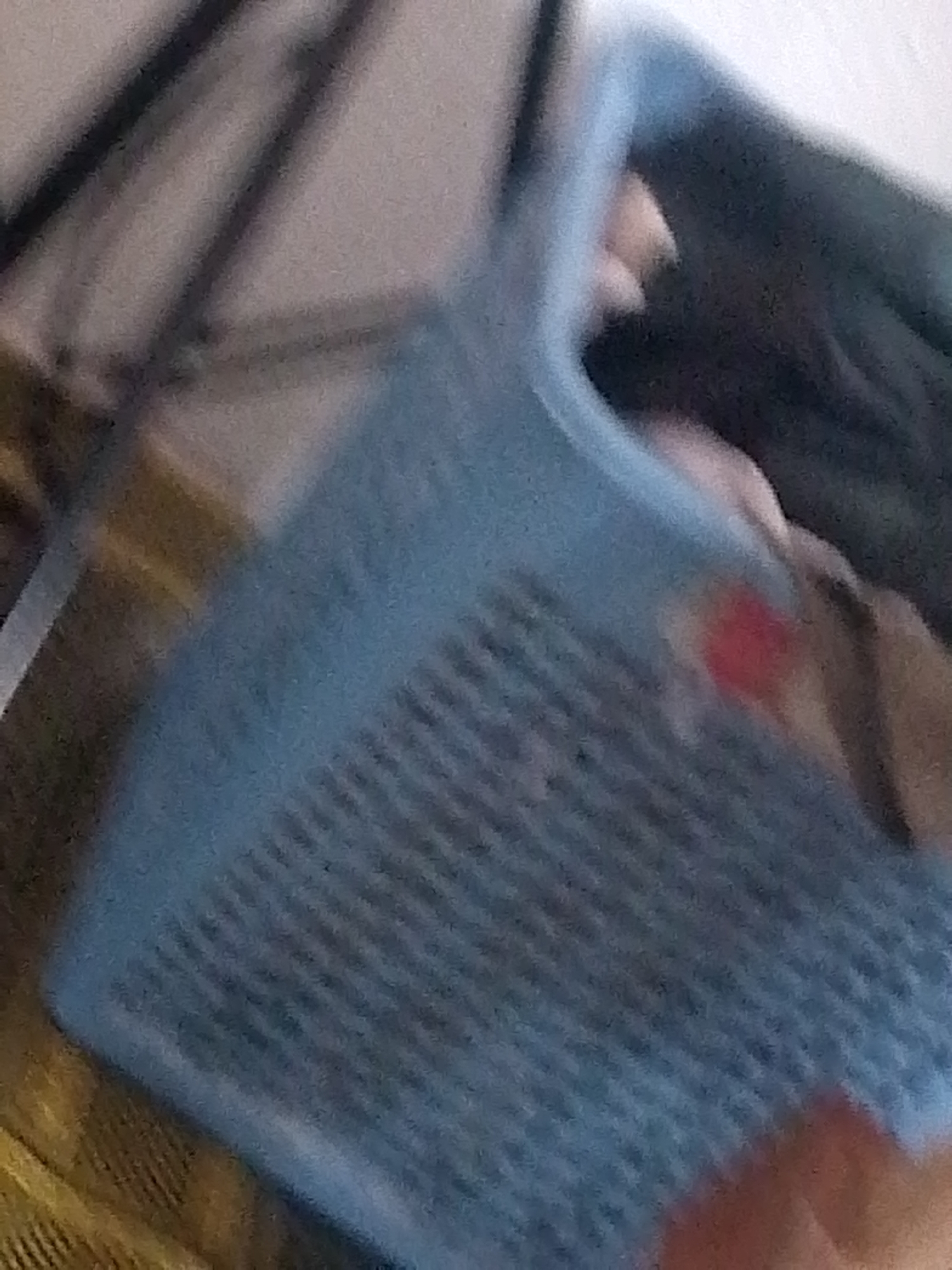 The image shows a close-up of a blue plastic basket with a woven texture. A person's hand, with a small red mark on the skin, is visible in the lower right portion of the frame, holding or touching the basket. The background is blurred and appears to be indoors, with some dark and light-colored fabric or surfaces visible. The scene is not specific to Jauro Yinu, Nigeria, or any identifiable urban location.