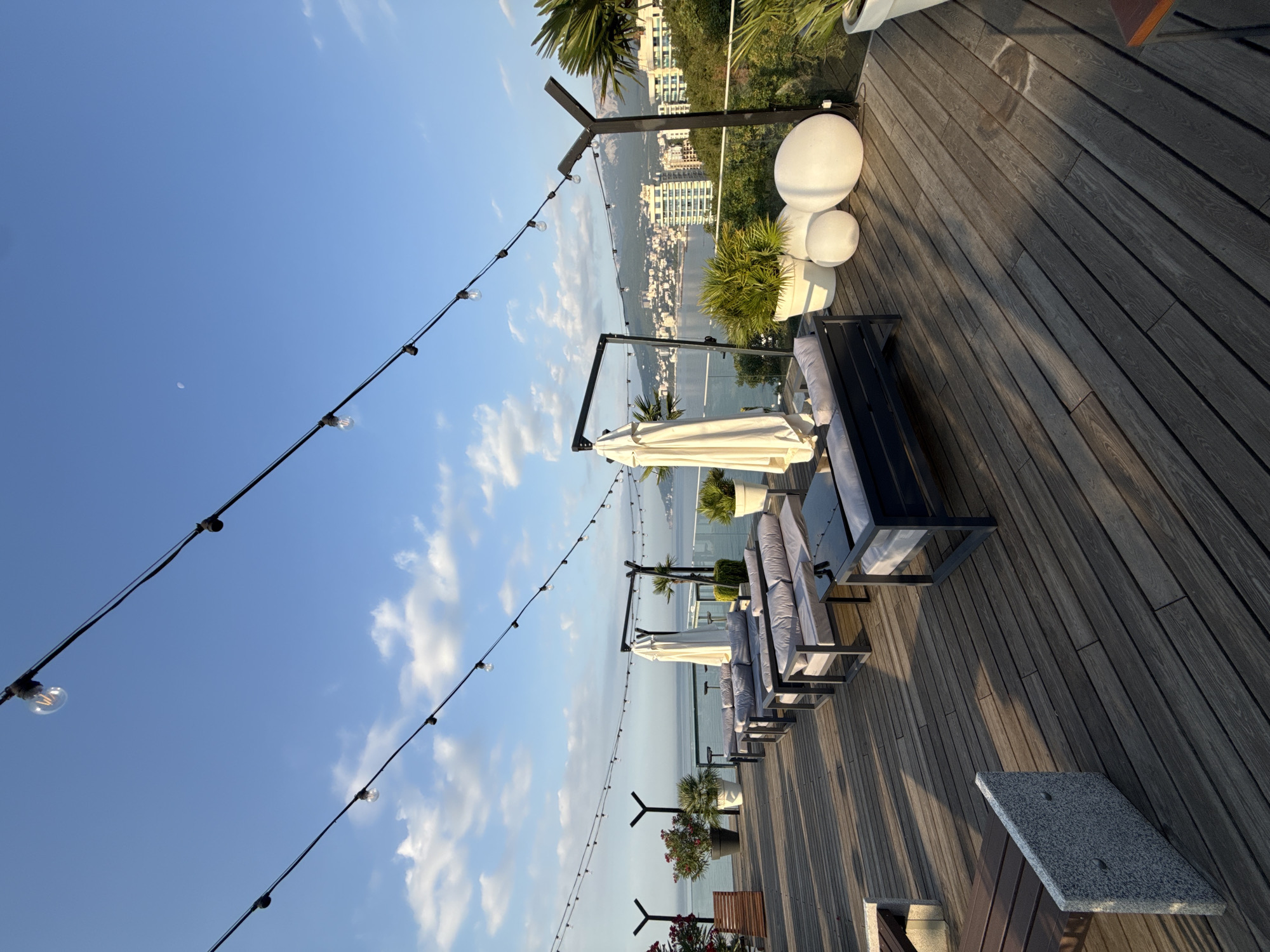 The image depicts an outdoor rooftop seating area overlooking the city of Yalta, Ukraine. The foreground is dominated by a wooden deck with rows of modern gray sofas and accompanying tables. String lights with visible bulbs are suspended above the seating area.
In the middle ground, white patio umbrellas are set up, suggesting a place for shade. Potted plants, including palm trees and flowering bushes, are strategically placed around the seating. To the right, large white spherical decorations add to the ambiance.
The background reveals a panoramic view of Yalta, with its distinctive architecture and buildings stretching along the coastline. The Black Sea is visible, with mountains forming a backdrop to the city. The sky is a clear blue with scattered clouds, and a faint crescent moon is visible. The lighting suggests it is either early morning or late afternoon, with long shadows cast across the deck. No people are present in the image.