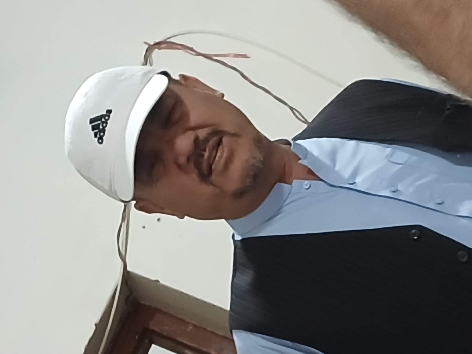 The image depicts a man indoors, likely in Quetta City Tehsil, Pakistan. He is shown from the chest up, with his head tilted slightly. He wears a white baseball cap with the black Adidas logo prominently displayed on the front. His eyes appear closed or are nearly shut, suggesting he might be resting or was captured in a moment of relaxation. He has a mustache and a short beard or stubble.
The man is dressed in a light blue collared shirt worn under a dark-colored vest or waistcoat with a subtle pinstripe pattern. The background consists of a plain, light-colored wall or ceiling. Several loose wires, one reddish-brown and one darker grey/brown, hang somewhat haphazardly against this background. In the lower part of the frame, a segment of a wooden structure, possibly a window or door frame, is visible. The lighting is soft and ambient, characteristic of an indoor setting with no direct bright sunlight. No specific activity other than possibly resting is discernible. The overall impression is a candid, unposed shot.