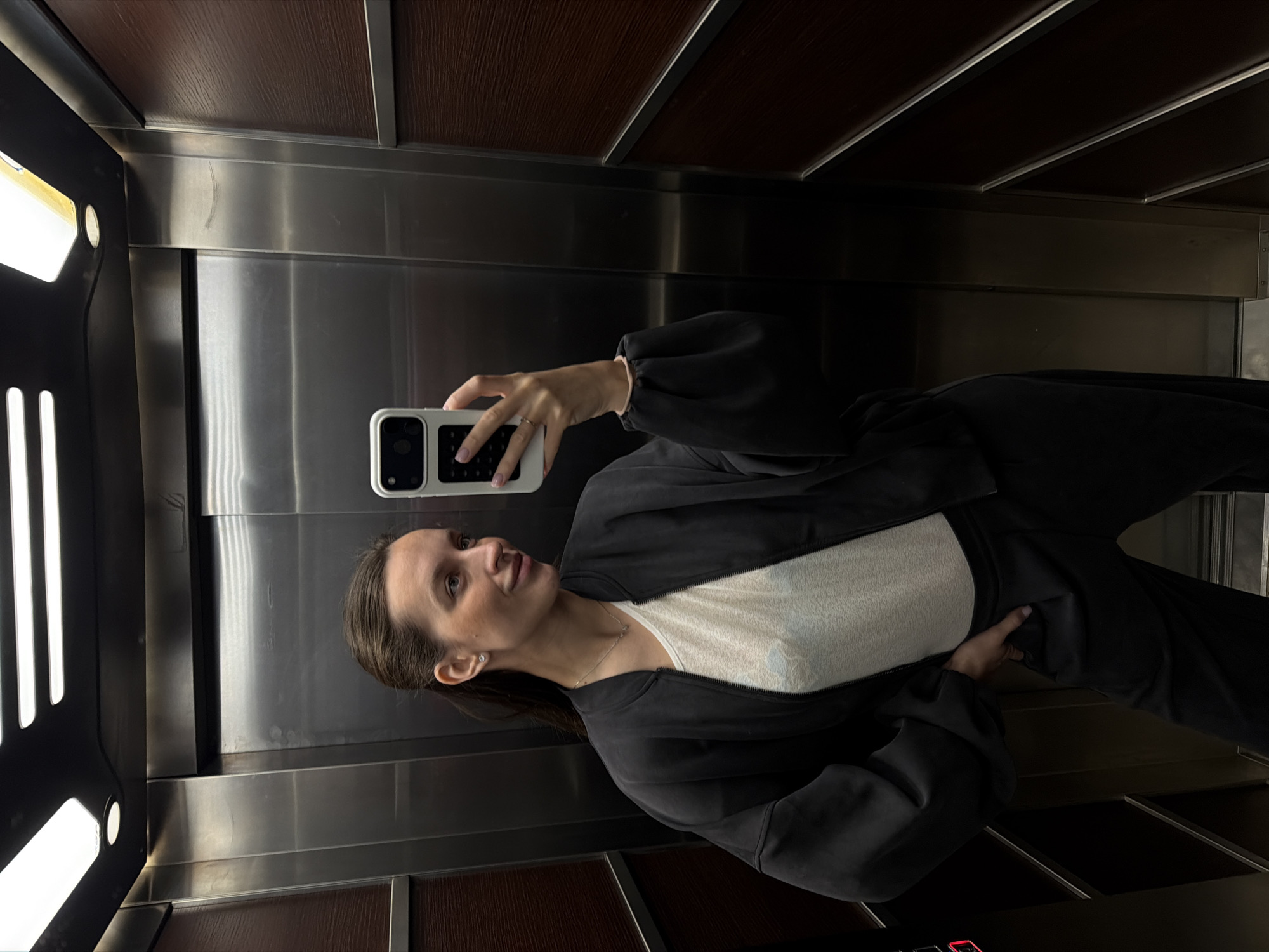 The image shows a woman taking a selfie in an elevator. The elevator has stainless steel walls and wood paneling on the upper and lower sections. There is a light fixture on the left side of the frame. The woman is holding a white smartphone and looking at the screen. She is wearing a white top and a black jacket with black pants. The elevator floor has buttons and indicators. The location is an elevator in Budva, Montenegro.