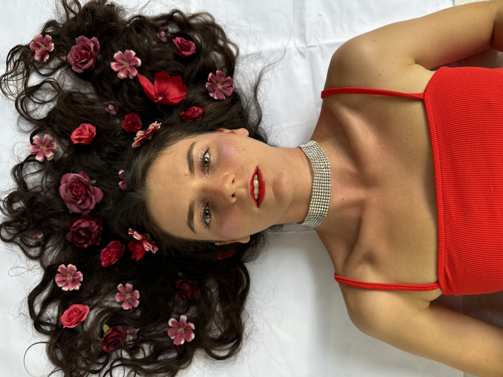 A woman lies on a white surface with her head and hair spread out. Her dark, wavy hair is adorned with several artificial flowers in shades of pink and red. She is wearing a red sleeveless top and a sparkling silver choker necklace. Her makeup includes pink blush and red lipstick. The scene does not contain any elements that would indicate a specific location in Mũi Né, Vietnam.