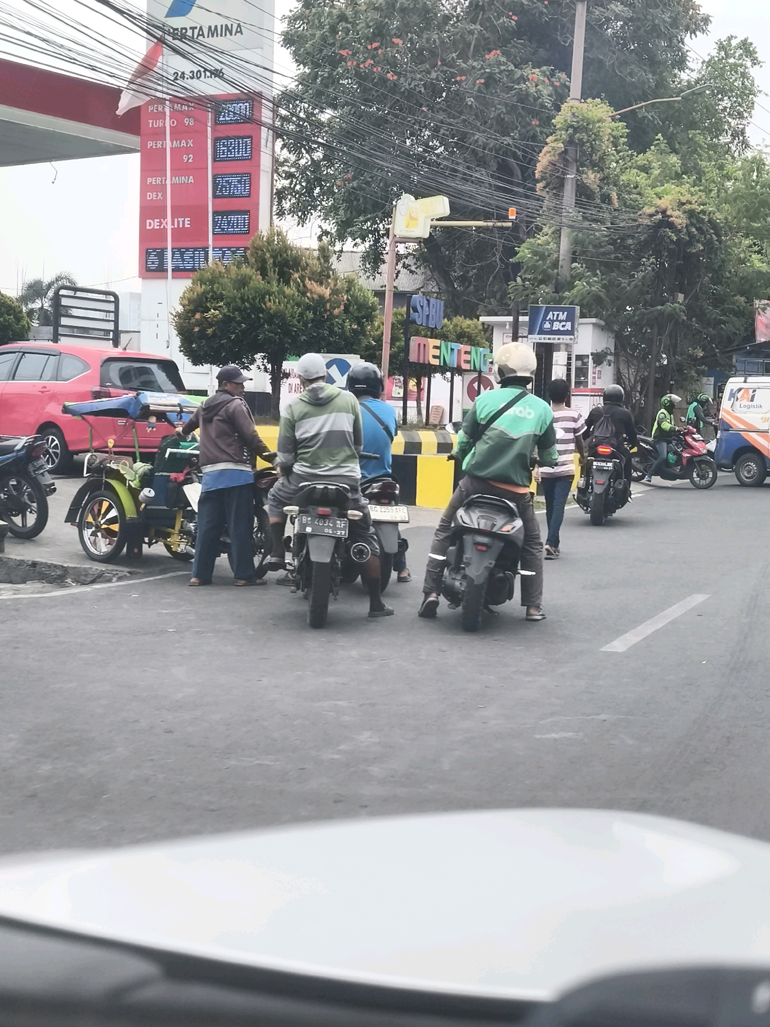 In Palembang, Indonesia, a street scene unfolds at a Pertamina gas station. A prominent red and white Pertamina sign displays fuel prices: Pertamax Turbo at 14,301.76, Pertamax 92 at 13,250, Pertamina Dex at 14,250, and Dexlite at 13,800. Several individuals on motorcycles are present. One person in a green jacket with "Gojek" visible is on a scooter. Another person in a grey and green jacket is also on a motorcycle, as is someone in a blue shirt. A man in a brown jacket and blue pants stands near a yellow and green motorized rickshaw. A red car is parked behind the rickshaw. In the background, an ATM BCA sign is visible, along with trees and utility wires overhead. The overall environment suggests a bustling urban setting.