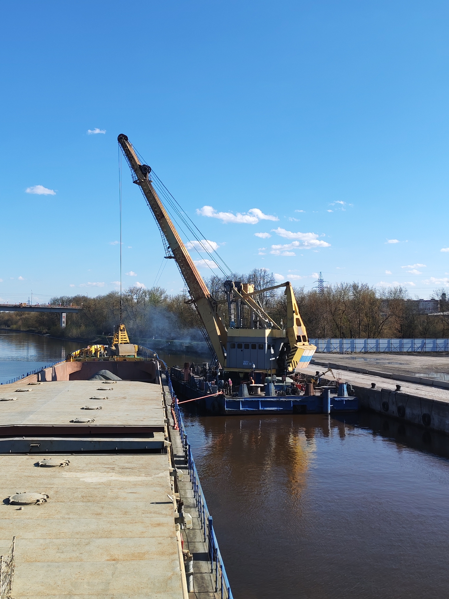 The image shows a scene at the Yakhroma River in Yakhroma, Russia. A large, yellow crane is positioned on a floating barge beside a dock and is loading material from another barge. The barge being loaded appears to contain gravel or similar material. Smoke rises from the area where the crane is operating. The water of the river is brownish in color. In the background, trees and a bridge are visible under a clear, blue sky with scattered clouds. A blue railing runs along the side of the barge where the image is taken. The photograph is taken from a high angle, looking down onto the scene.