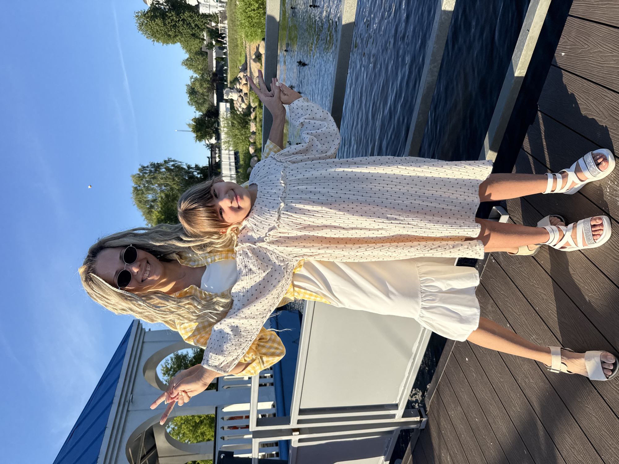 An adult female and a young female stand on a dark brown wooden deck, posing for a photograph under clear blue skies in Dumino, Russia. The adult female, positioned slightly behind and to the left of the younger female, has blonde dreadlock-style hair, wears dark oval sunglasses, a yellow and white checkered shirt over a white top, a white tiered dress, and white sandals. She forms a peace sign with her left hand and has her right arm extended behind the younger female. The young female, with brown hair and bangs, wears a long-sleeved white dress with a dark dotted pattern and white sandals. She forms a peace sign with her left hand and holds a silver metal railing with her right hand. Both individuals are smiling directly at the camera.
The metal railing separates the deck from a body of water, where several ducks are visible in the midground. In the left background, a white structure with a blue gabled roof and arched openings is partially visible. The background also includes green trees, shrubs, and a landscaped area with large rocks across the water. Further in the distance, white buildings, including one with a visible dome, are present. The scene is brightly illuminated by sunlight, casting distinct shadows on the wooden deck.