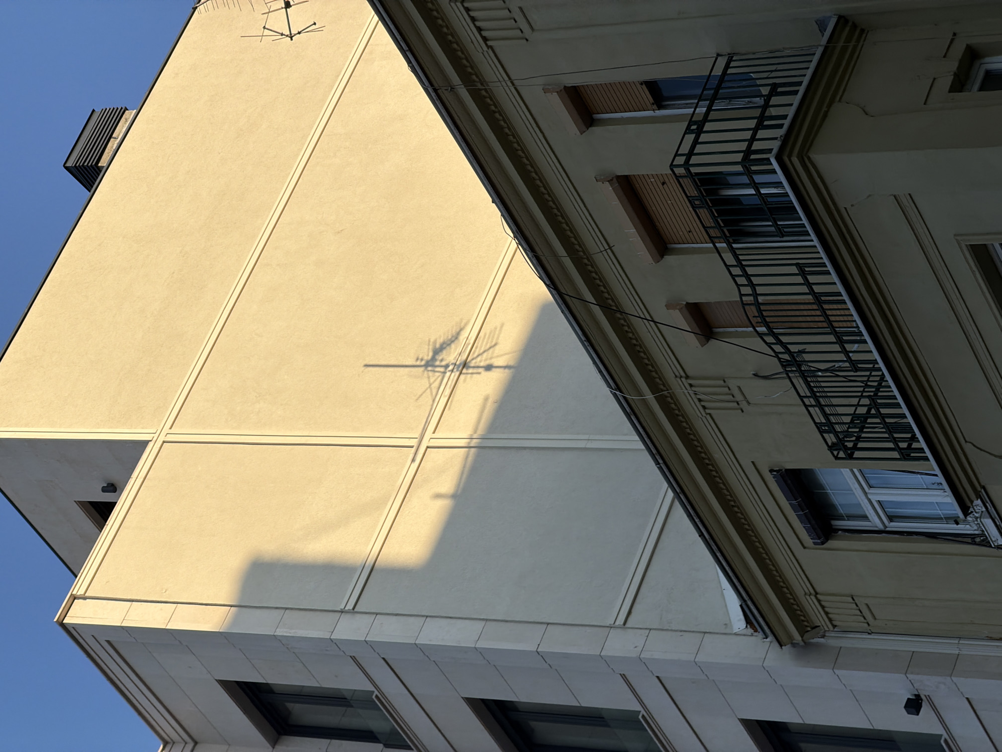 The image shows two buildings in Belgrade, Serbia, viewed from a low angle. The building on the left is modern, with a flat, light-colored facade divided into rectangular sections. A shadow of an antenna is cast on the facade. The building on the right is older, with more ornate architectural details, including a balcony with a black metal railing. Windows with closed shutters are visible. The sky is blue.