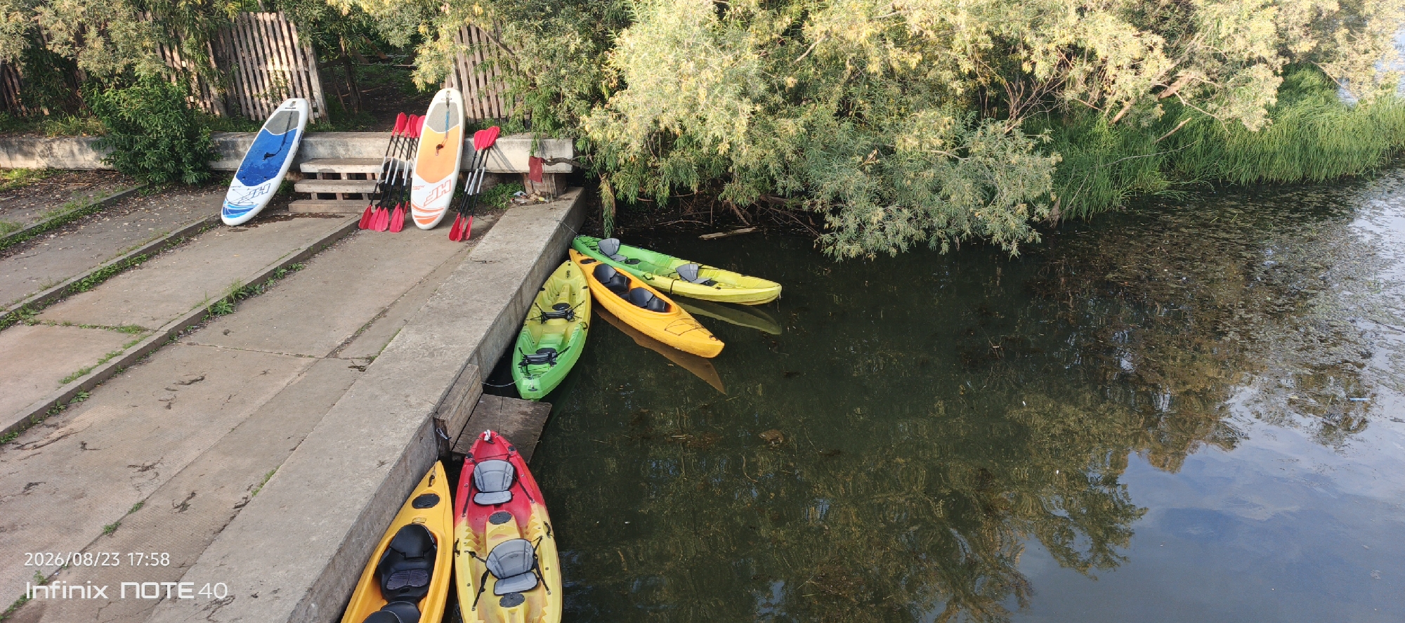 The image depicts an outdoor scene beside a body of water, likely a river or lake, in Surgut, Russia. A concrete embankment lines the water's edge. On the embankment, several kayaks are positioned, with two yellow kayaks and two green kayaks visible. One yellow kayak and one red and yellow kayak are closer to the viewer, while two green kayaks and one yellow kayak are further away, some partially submerged in the water. To the left of the kayaks, on the concrete, are two stand-up paddleboards, one white and blue, and another white. Several red paddles are leaning against the stand-up paddleboards. Behind the embankment, a wooden fence and dense green foliage are visible, suggesting a natural or park-like setting. The water surface is dark and reflects the surrounding trees. The timestamp "2026/08/23 17:58" and "Infinix NOTE 40" are visible in the bottom left corner of the image. There are no people visible in the scene.