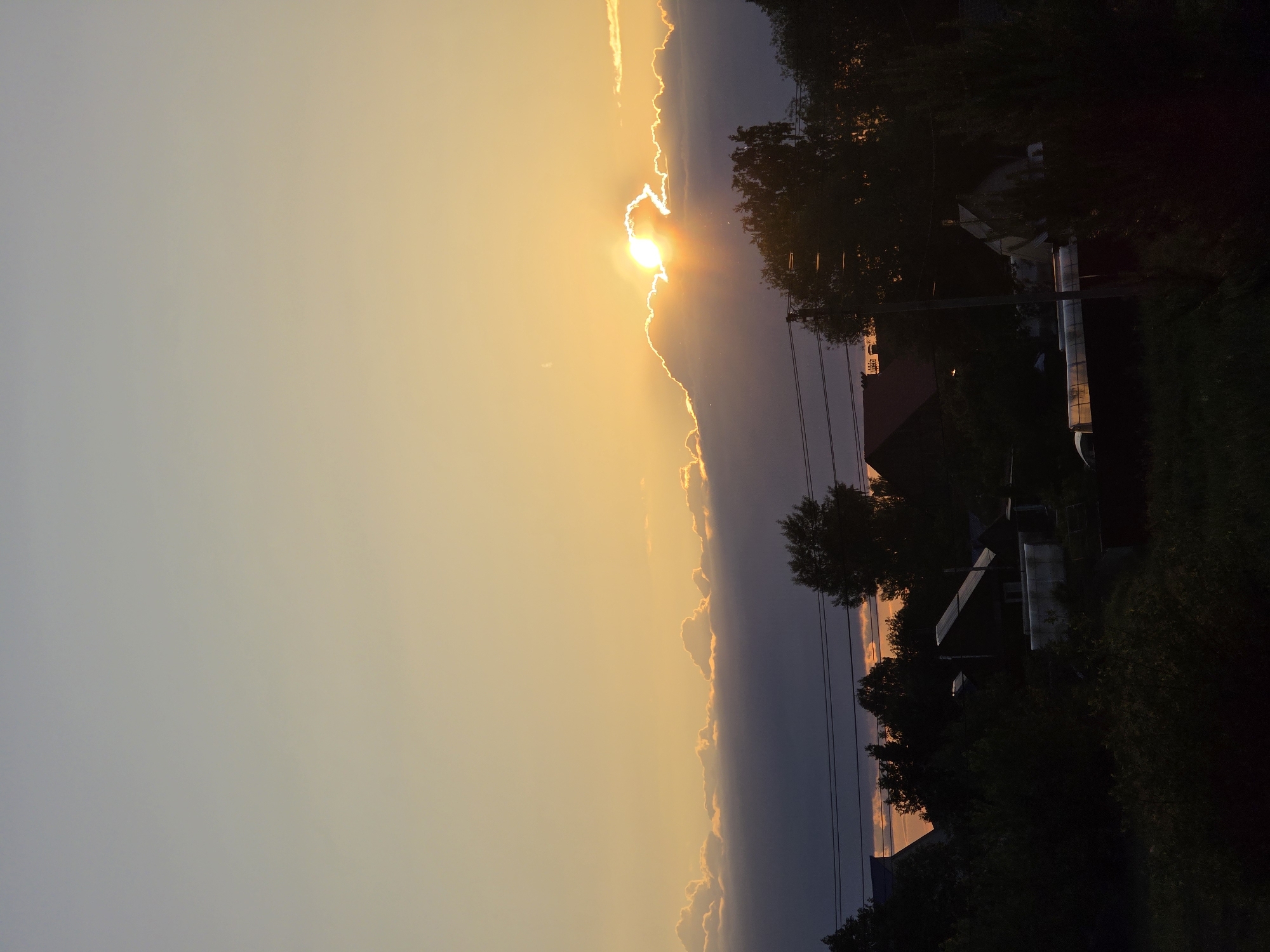 The image, presented in portrait orientation, captures a vibrant sunset scene over a rural or suburban area in Chaikovsky, Russia. The majority of the frame, from the left edge towards the center-right, is dominated by the sky, displaying a gradient from a pale gray-blue on the far left to a warm, brilliant orange and yellow closer to the center-right.
A prominent, elongated cloud formation stretches from the top to the bottom of the image, roughly horizontally across the depicted landscape. The sun, a bright luminous disc, is partially obscured behind these clouds towards the center-right, casting an intense golden glow that outlines the cloud edges and creates a starburst effect.
The right side of the image, which would be the bottom of the landscape if rotated to a horizontal view, is largely composed of a dark, silhouetted foreground. This area features dense masses of trees and foliage, particularly in the lower-right corner. Through the shadows, several buildings, possibly houses, are faintly discernible, with some roofs exhibiting a subtle reddish-orange reflection from the sky. Utility poles and a network of overhead power lines are clearly visible running across the mid-right section, adding a structural element to the shadowy foreground.
No people or explicit activities are depicted. There is no visible text in the image. The time of day is clearly sunset, creating a dramatic and peaceful atmosphere.