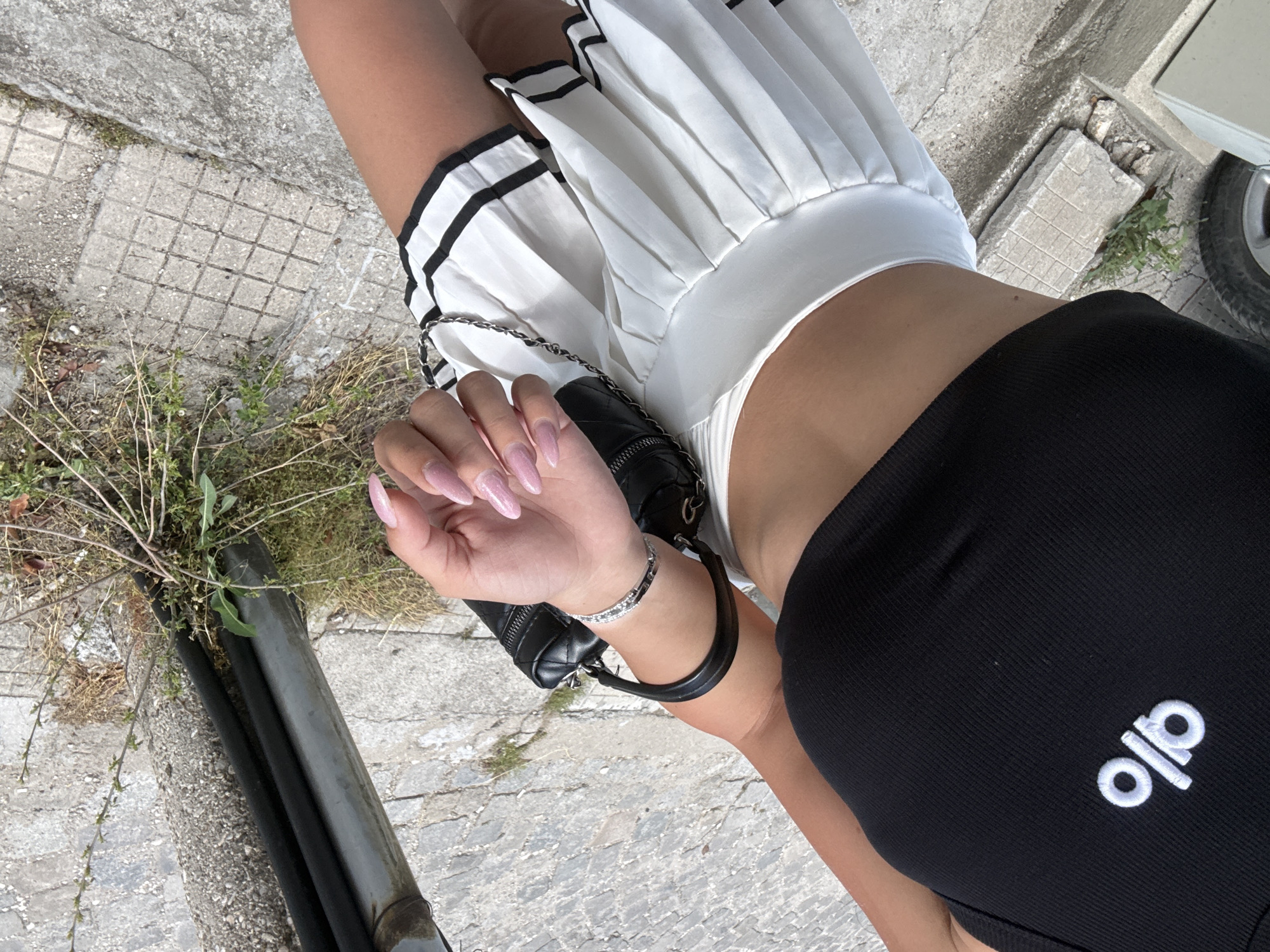 A close-up, slightly tilted, high-angle shot depicts a person wearing a black tank top with "ALO" in white lettering, a white pleated skirt with black trim, and jewelry. The person's left arm and hand are visible, holding a black purse with a silver chain. A silver bracelet is on the wrist. The person has light skin, and their fingernails are painted with a light pink color.
The background consists of a concrete surface with cracks and grid patterns, a metal pipe, and some weeds growing along the edge. A partial view of a car tire is visible in the upper right corner.