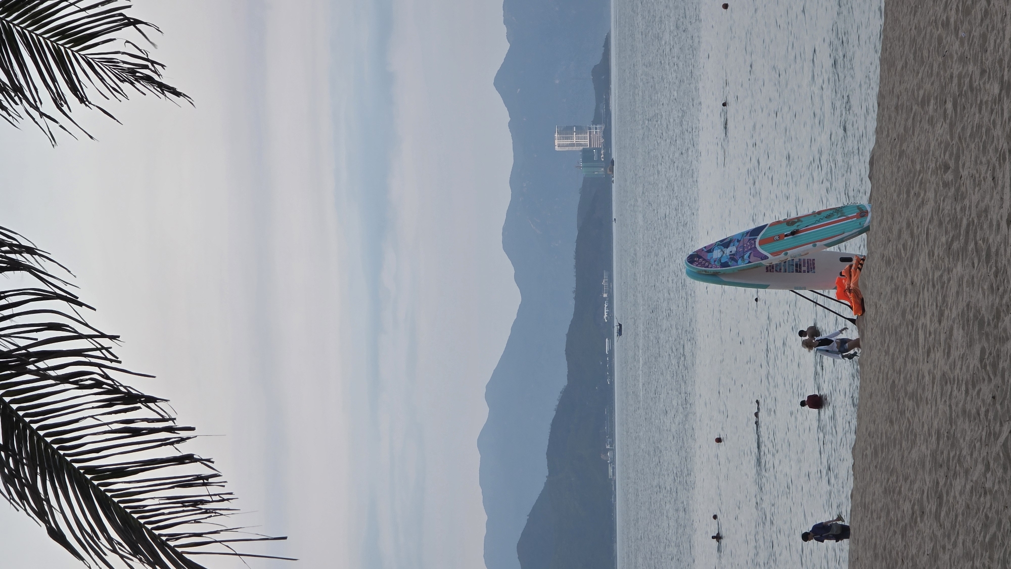 The image captures a serene beach scene, likely on an overcast or hazy day, with palm fronds framing the left side. The sky, occupying the upper portion, is a soft, uniform light grey-blue. In the mid-ground, a vast expanse of calm, greyish-blue ocean stretches horizontally, with gentle ripples on its surface and several small, dark buoys floating at various distances.
Along the right edge of the image, a sandy beach forms the foreground. Two brightly colored paddleboards are prominently displayed near the water's edge, leaning upright on stands. One board is light blue with vibrant, colorful patterns, while the other is predominantly white. An orange life vest is draped over the white paddleboard.
Several people are visible in and near the water. One individual, dressed in a light top and dark bottoms, stands on the sandy shore close to the white paddleboard. Further out in the ocean, a few other people are seen wading or swimming, their forms partially submerged. One person wears a red top.
In the background, layers of hazy blue mountains rise from the distant coastline, creating a sense of depth and vastness. A large, multi-story building with a distinctive white facade is visible on the distant shore, nestled against the mountains, suggesting a developed coastal area. The overall atmosphere is peaceful and relaxed. Some text or branding is visible on the paddleboards, but it is not clearly legible. Given the context, this is a typical scene from Nha Trang, Vietnam.