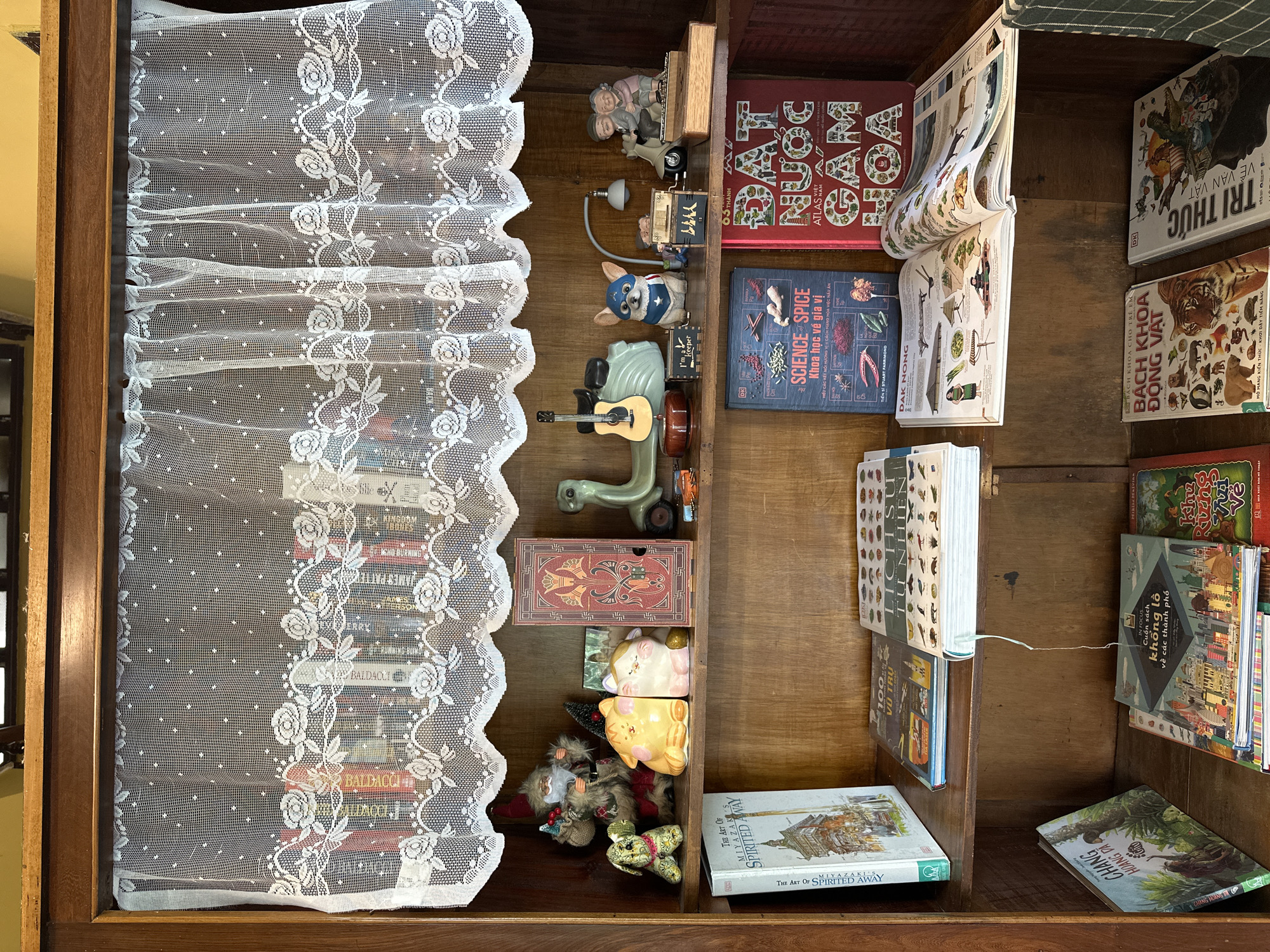The image depicts a wooden bookshelf filled with books and decorative items. The bookshelf is likely located in Da Lat, Vietnam, as suggested by the presence of Vietnamese language books on the shelves, such as "Đất Nước Cẩm Hoa" and "Bách Khoa Động Vật."
On the top shelf, behind a white lace curtain, a collection of books with colorful spines is visible. In front of the books, various decorative items are displayed. These include a Santa Claus figurine, a stuffed dog, two ceramic cat figurines, a book with an ornate cover, a green toy scooter with a guitar on it, a small lamp, a dog figurine wearing a superhero cape, a small toy figure, and a figurine of an elderly couple riding a scooter.
The middle shelf contains more books. On the left, a book titled "The Art of Spirited Away" is prominently displayed. Next to it are other books, one with "100 VÙ" on its spine and another with "LỊCH SỬ TỰ NHIÊN" visible. An open book with illustrations of food and people is laid flat in the center of the shelf.
The bottom shelf also holds a variety of books. On the left, a book with a jungle scene and the title "CHÂNG HOÀNG VÀ" is visible. To the right of that, an open book with the title "Cuốn Sách Khổng Lồ về Các Thành Phố" is displayed, along with other stacked books. Further to the right, two more books are visible: "BÁCH KHOA THẾ GIỚI ĐỘNG VẬT" and "TRI THỨC VỀ VẠN VẬT". The overall environment suggests a cozy and curated collection of knowledge and decorative items.