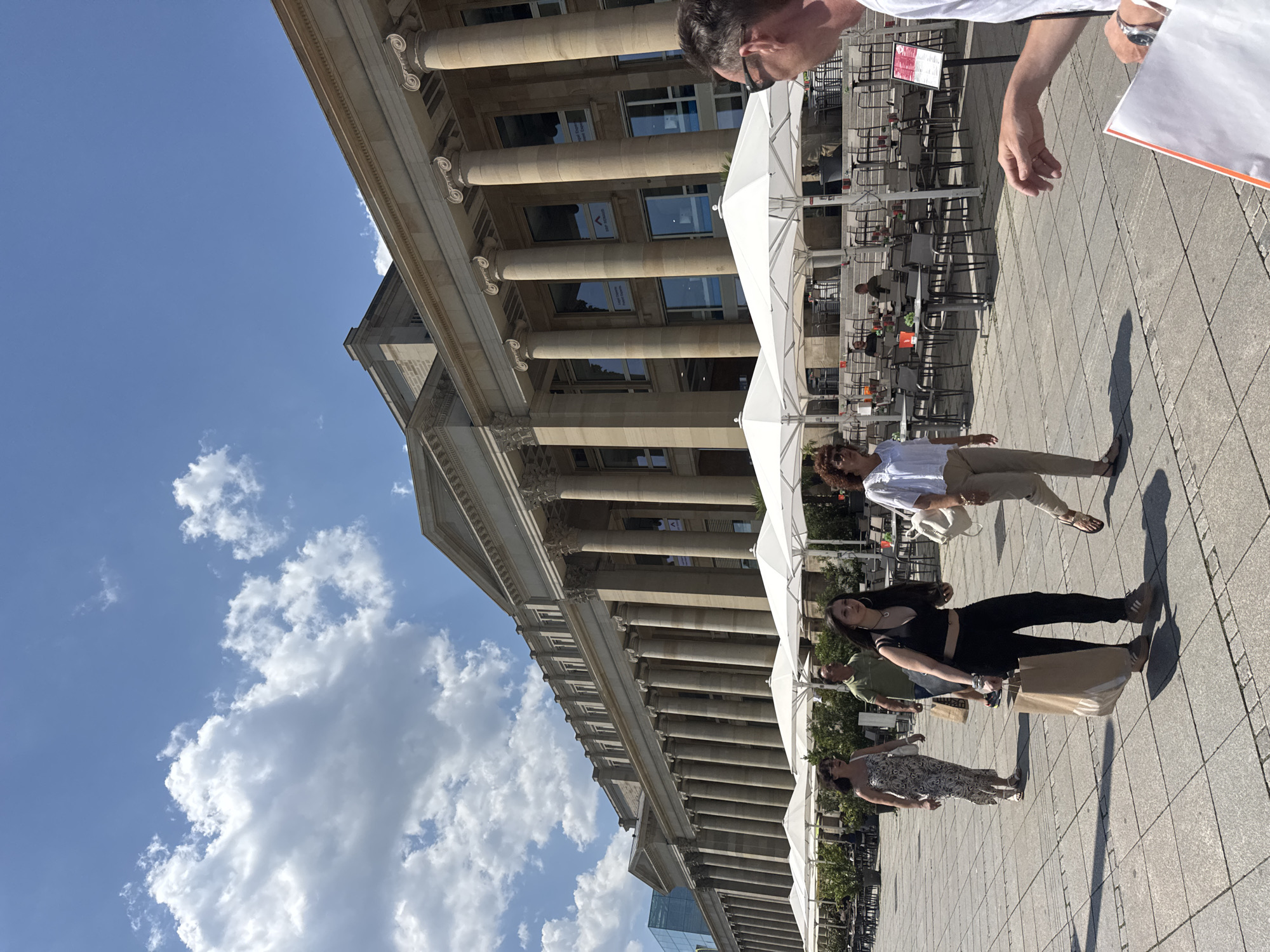 The image shows a street scene in Stuttgart, Germany, featuring the New Palace (Neues Schloss) on a sunny day with scattered clouds. The New Palace is a large Baroque building with a prominent colonnade. In the foreground, several people are walking on a paved plaza. A woman in a floral dress is walking from left to right, followed by a man in a green shirt. To their right, a young woman in a black top and pants is walking towards the camera, carrying a large paper shopping bag. Further to the right, a woman in a white top and tan pants is walking in the same direction as the young woman. Several white umbrellas and outdoor seating for a cafe are visible in the middle ground, with the New Palace serving as the backdrop. The image captures a moment of everyday life in a public space in Stuttgart.