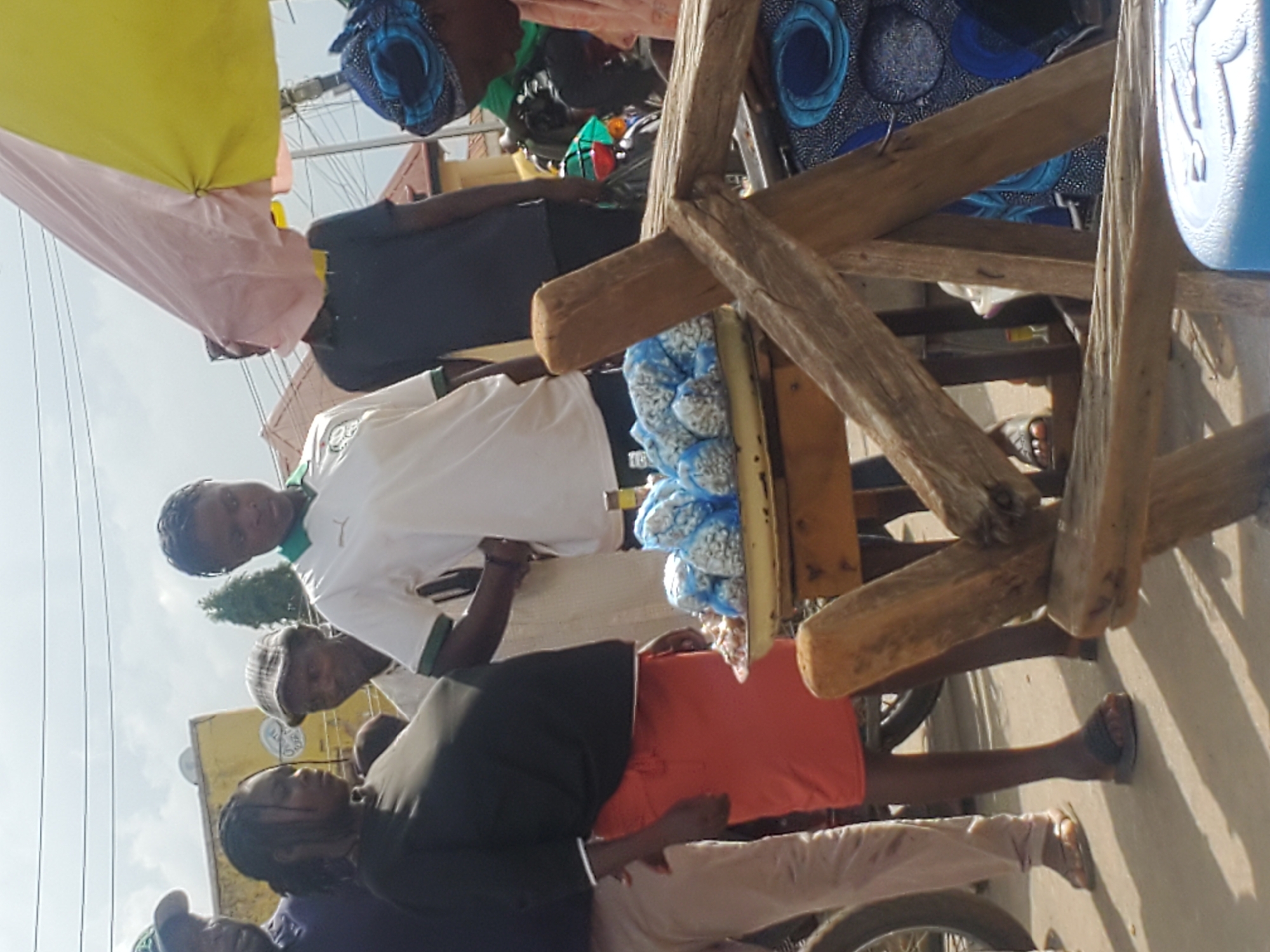 In a market scene in Gwong, Nigeria, a group of people are gathered around a wooden cart laden with goods. The cart has a yellow canopy and appears to be a stall for selling items. Several individuals, both male and female, are visible, some wearing traditional clothing and headwraps. One person, possibly a vendor, is wearing a white shirt with green trim and is handling some of the merchandise which is packaged in blue bags, resembling snacks or small produce. Another person is wearing a black top and an orange skirt. The overall setting suggests a bustling outdoor market, possibly during the day given the lighting. The people appear engaged in trade or interaction. There are no visible text elements in the image.