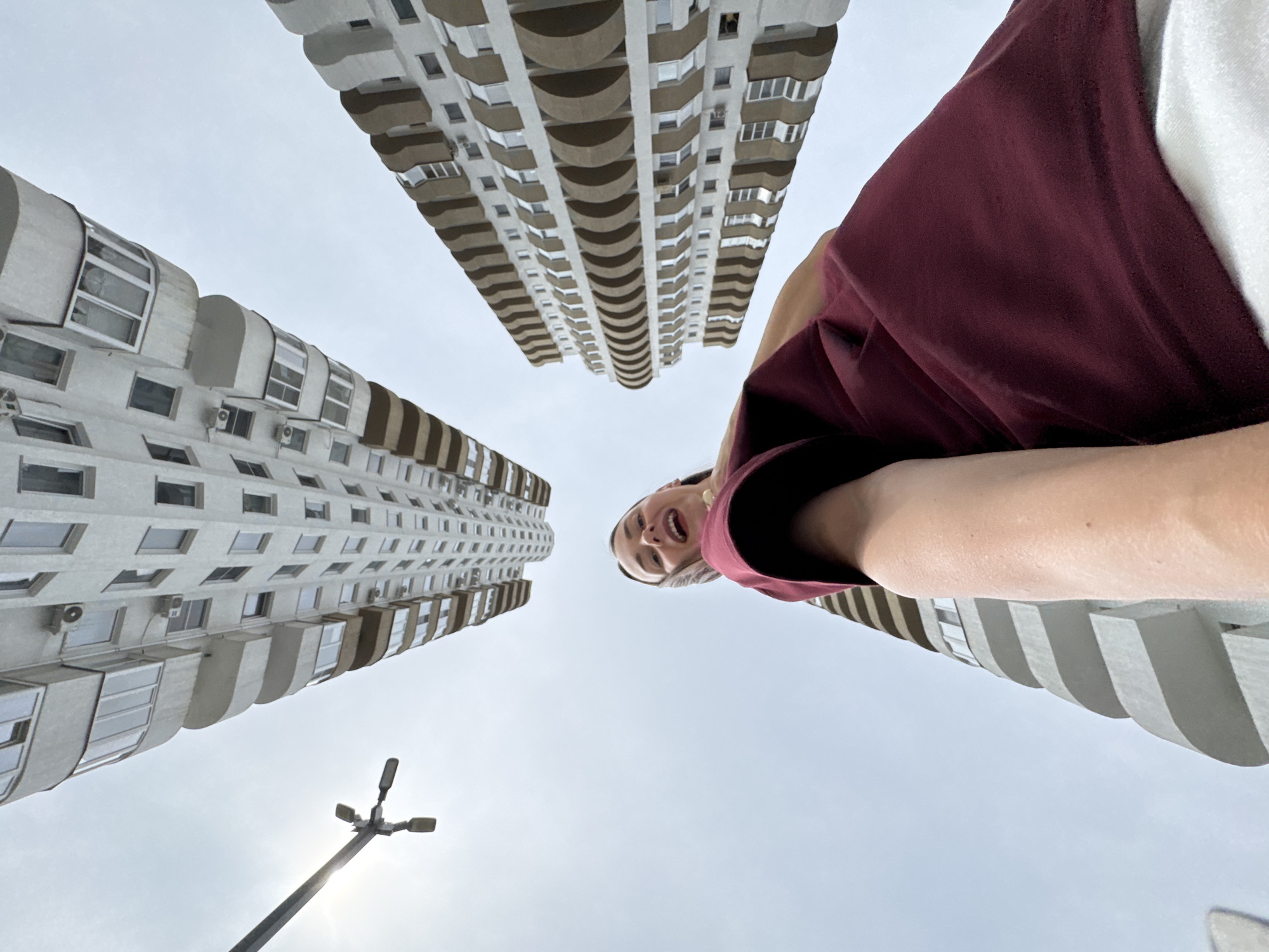 A low-angle shot captures a young woman holding a phone, taking a selfie, while standing between two tall, modern apartment buildings in Sochi, Russia. The buildings, with their white facades and curved balconies, tower above, reaching towards a slightly overcast sky. The woman is wearing a maroon t-shirt and white shorts. To the left, a street lamp is visible against the sky, with the sun peeking out from behind one of the buildings. The perspective creates a sense of being dwarfed by the urban architecture.