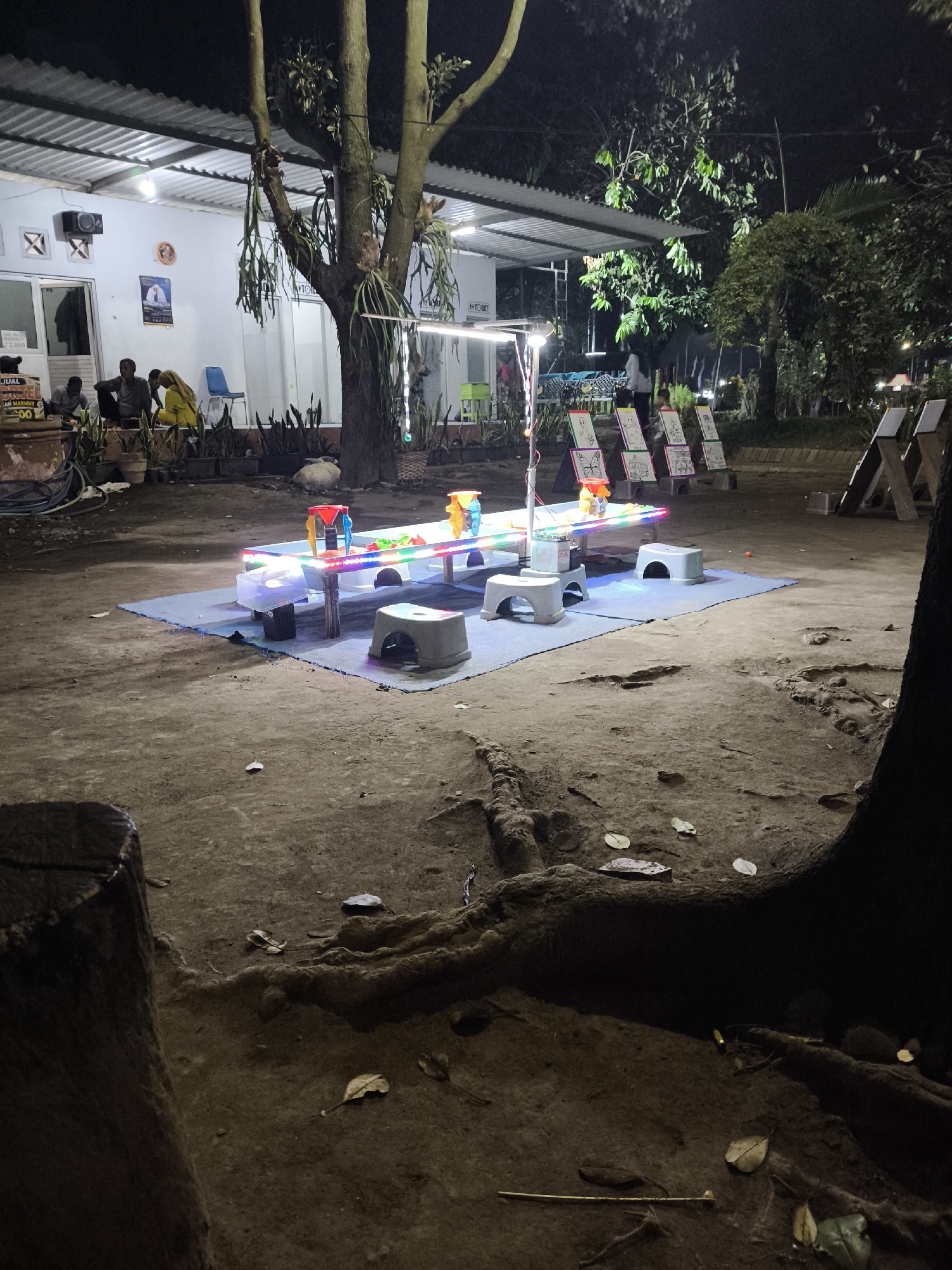 This is a nighttime photograph taken outdoors at a street-side food stall or community area in Lumajang, Indonesia. In the foreground, a tree stump and exposed tree roots are visible on the dark, sandy ground, scattered with dry leaves. The main subject is a low, illuminated table set up on a blue mat. On the table are various colorful plastic toys and objects, possibly part of a children's activity or game. Several small, grey plastic stools are placed around the mat.
In the mid-ground, a white building with a corrugated metal roof is visible, illuminated by overhead lights. Several people are seated or standing near the building. One man in a grey shirt is sitting on the ground. Another person, possibly a woman in a yellow shirt, is also seated. A blue plastic chair is visible. Signs are posted on the building, one with text that appears to be in Indonesian.
Further in the background, more trees and foliage are present, along with additional illuminated stands or tables displaying what appear to be drawings or artwork. The overall atmosphere suggests a casual, community gathering space illuminated by artificial lights in the evening. The presence of the building with the "TOILET" sign indicates a public facility.