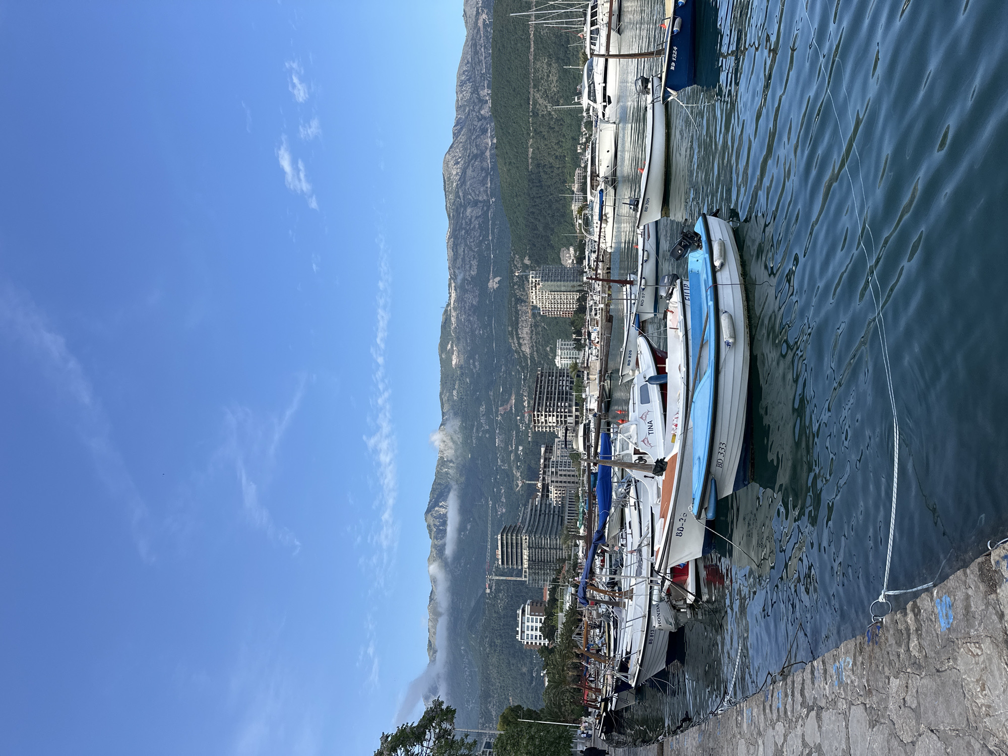 The image shows a port in Budva, Montenegro, with several boats moored along a stone pier. The water is calm and reflects the sky. The boats are of different sizes and colors, with some having registration numbers like "BD 813," "BD-25," "BD 333," "BD 826," "BD 57," "BD 708," and "BD 1324." The side of one boat has the word "HONDA," while another has "TINA." In the background, there is a view of buildings and mountains, partly obscured by clouds. The sky is blue with scattered clouds. A crane is visible among the buildings, indicating ongoing construction. A few trees are visible on the lower left of the image.