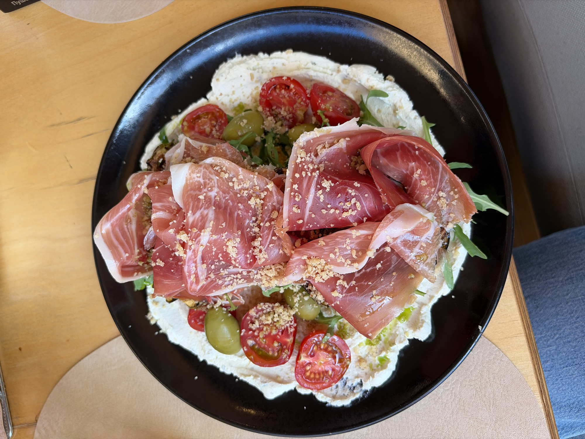 A black, oval-shaped plate, positioned on a light brown wooden table, is filled with a culinary arrangement. A thick, off-white creamy substance forms an uneven base layer. Layered on top are multiple slices of reddish-pink cured ham, exhibiting white fat marbling, with some slices folded. Scattered across the ham and other components are light-colored crumbs. Several halved red cherry tomatoes and whole green olives are distributed throughout the dish. Small amounts of dark green leafy greens are visible beneath the primary ingredients, and a drizzle of green oil is present on the creamy base. To the left of the plate, a lighter brown placemat is partially visible. In the lower-left corner, a segment of a leg clad in blue denim jeans is present. The upper right background reveals a sliver of a dark object with partially visible white Cyrillic text, including the character "П". The scene is illuminated by bright ambient light. The location is in the city of Unknown, Unknown.