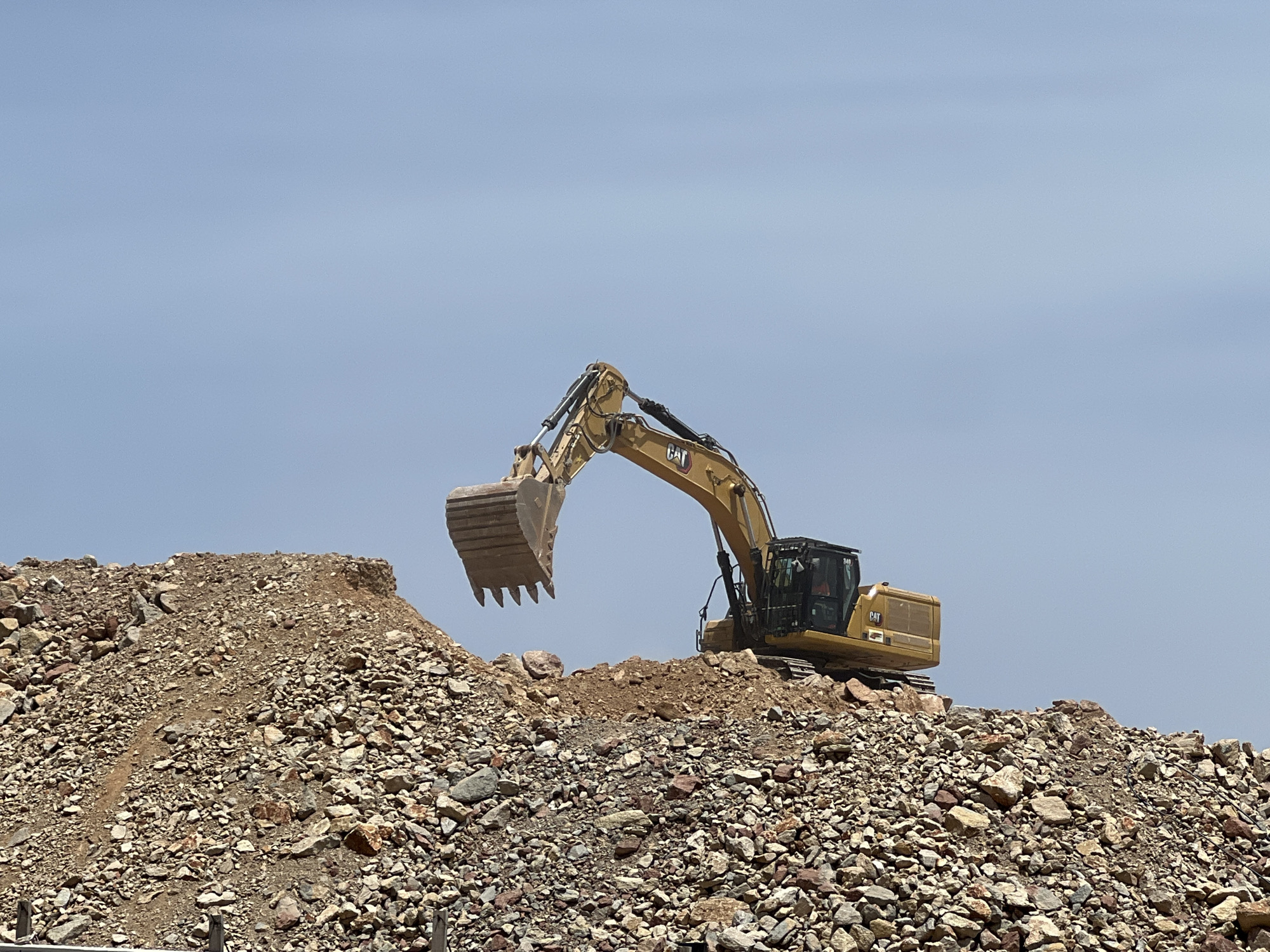 A yellow Caterpillar excavator is operating on a rocky incline under a clear, pale blue sky. The excavator, with the CAT logo clearly visible on its side, is positioned on a pile of brown and grey rocks and dirt. Its articulated arm is raised, and its bucket, also yellow and showing signs of use, is poised as if to scoop more material. The cab of the excavator is visible, and it appears a person is inside, operating the machinery. The scene suggests a construction or excavation project is underway. The lighting indicates it is daytime, likely late morning or early afternoon, with no clouds visible. The arid, rocky terrain is consistent with the landscape of Gibraltar.