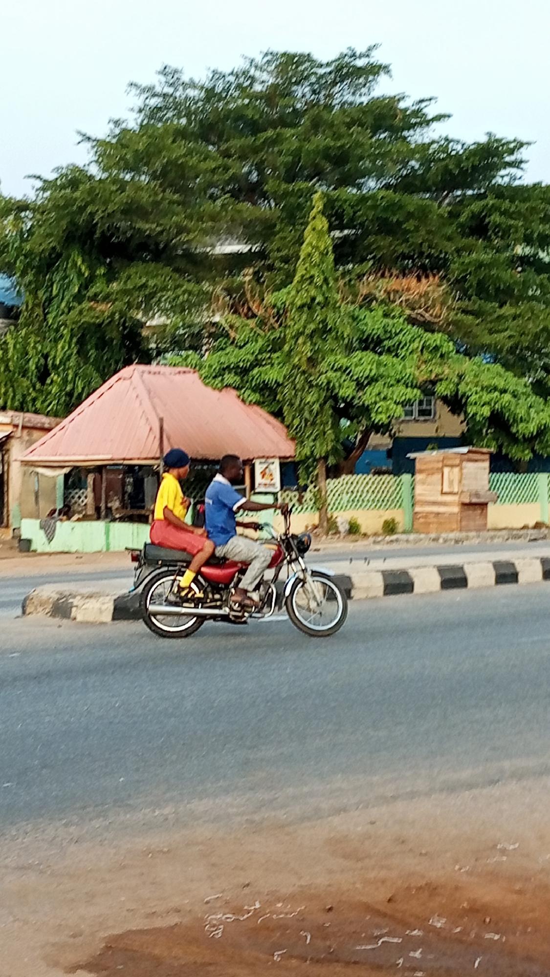 In Adankolo, Nigeria, two people ride a red motorcycle on a paved road. The driver, a man, wears a blue shirt and light-colored pants. His passenger, also a man, wears a yellow shirt, red pants, and a blue hat. Buildings and trees are visible in the background.