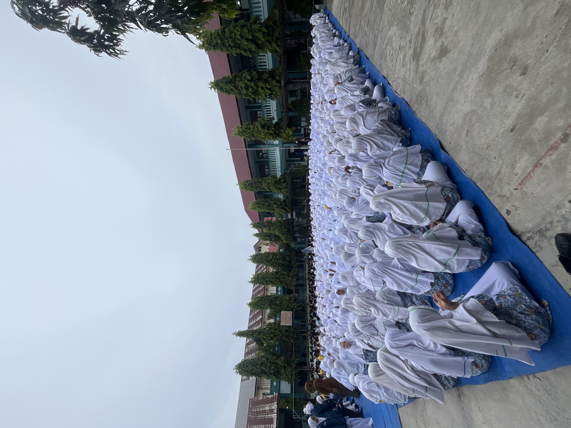 An aerial perspective shows a large gathering of individuals, predominantly young people, seated on a vibrant blue tarp spread across a paved courtyard. The individuals are uniformly dressed in white Islamic attire, consisting of headscarves and long garments, many with decorative patterns. A significant portion of the group appears to be female, based on the attire. They are arranged in neat rows, facing forward, suggesting a communal event or activity.
In the background, a two-story building with a red-tiled roof and light green exterior walls stands prominently. The building features a row of windows and balconies. Several tall, mature trees with dark green foliage are strategically planted in front of the building, creating a natural backdrop. The sky above is overcast and light gray, indicating diffused daylight. The ground surface is a mixture of concrete and the blue tarp, with some faint red markings visible on the concrete.
On the left side of the image, a few individuals are standing, one wearing a brown hijab and a beige garment, and others in dark blue jackets. To the far left, a glimpse of a white building is visible. The overall scene suggests an outdoor assembly at an educational institution or community center in Lhokseumawe, Indonesia, likely during a school event or religious observance.