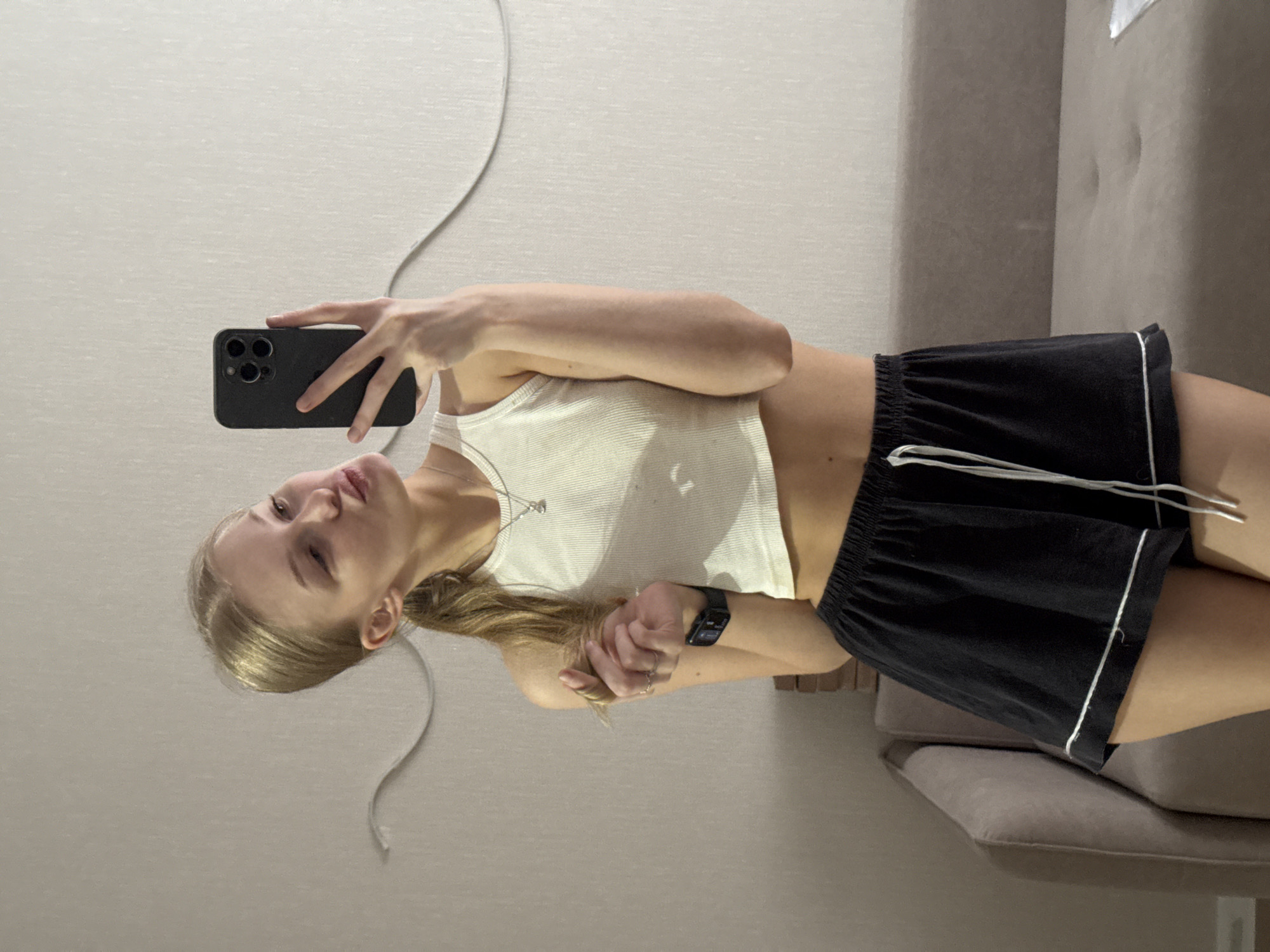 A young Caucasian woman with blonde hair pulled into a ponytail and wearing a white crop top and black shorts is taking a selfie in what appears to be a residential interior, possibly an apartment in Perm, Russia. She is holding a black smartphone in her right hand, angled to capture her reflection. Her left hand is near her face, playing with her hair, and she is making a kissing face. She is wearing a watch on her left wrist and a necklace. In the background, a muted beige couch is visible, and the wall has a subtle wavy decorative element. The lighting suggests an indoor setting, possibly during the day or evening, with no specific city landmarks clearly identifiable.