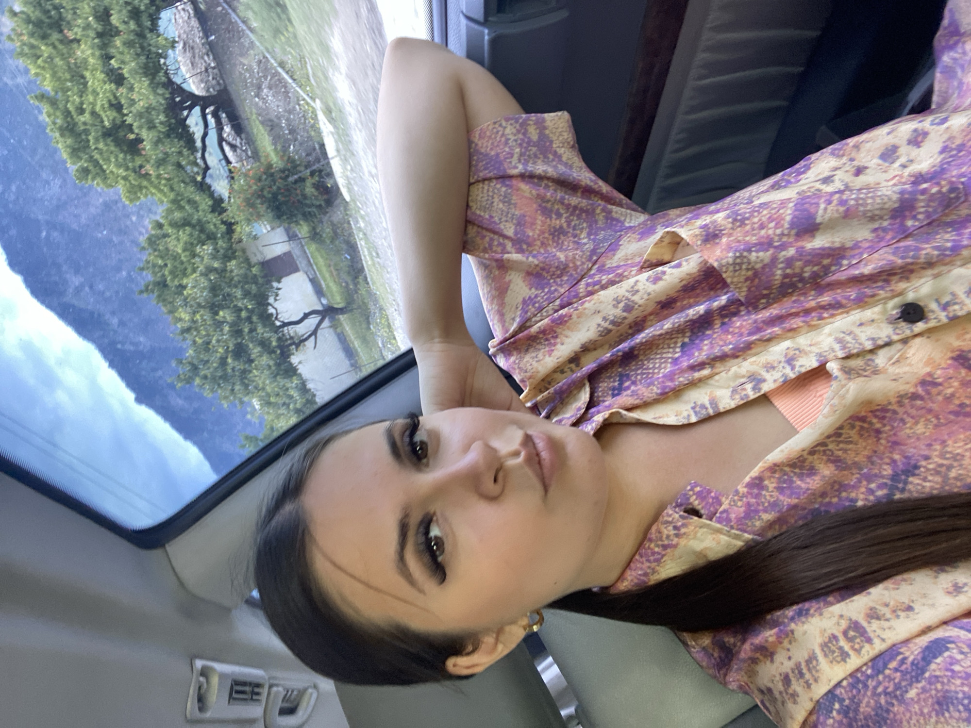A woman is seen from the chest up, sitting inside a vehicle. She is wearing a light-colored, patterned button-up shirt with purple and orange accents, and a peach-colored tank top underneath. Her left arm is bent, resting on her raised left elbow, with her hand behind her head. The woman has long dark hair pulled back into a ponytail, prominent false eyelashes, and is looking towards the camera with her lips slightly pursed. The background, visible through the vehicle's window, shows a mountainous landscape with green trees and a clear blue sky. The interior of the vehicle is also visible, including a headrest and a part of the dashboard. The image was taken in the city of Irganai, Russia.