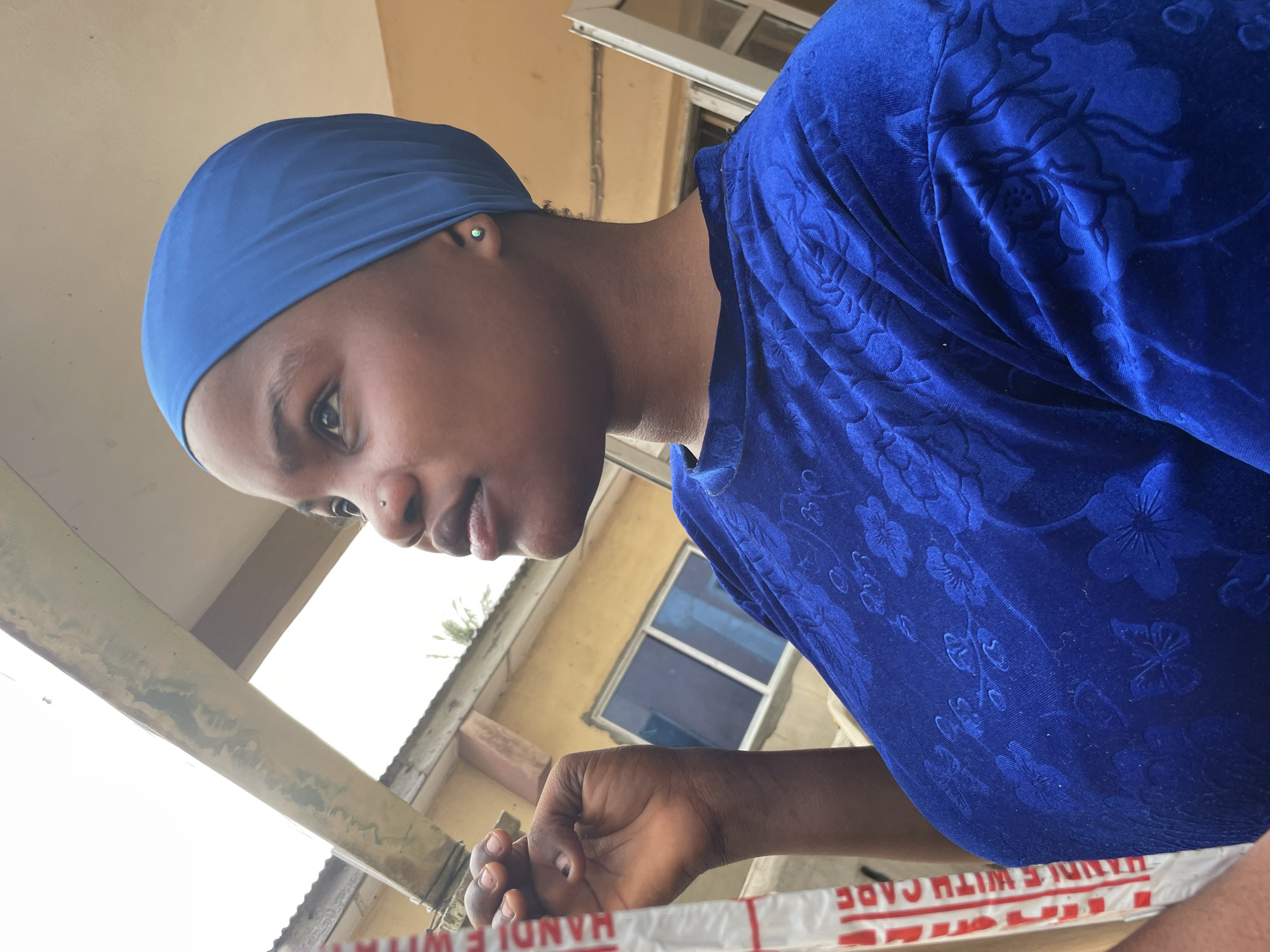 A medium, upward-angle shot shows a dark-skinned person wearing a blue head wrap and a blue, patterned velvet top. The person has a nose piercing and is looking downward and to the left. The hand of the person is visible in the foreground. The background includes a partial view of a light-colored building with a window, a corrugated metal roof, and a structural support beam. A white box with red lettering reading "HANDLE WITH CARE" is partially visible at the bottom of the frame. Based on the architectural style and the presence of "HANDLE WITH CARE" packaging, the image was likely taken in an urban environment in Badagry, Nigeria.
