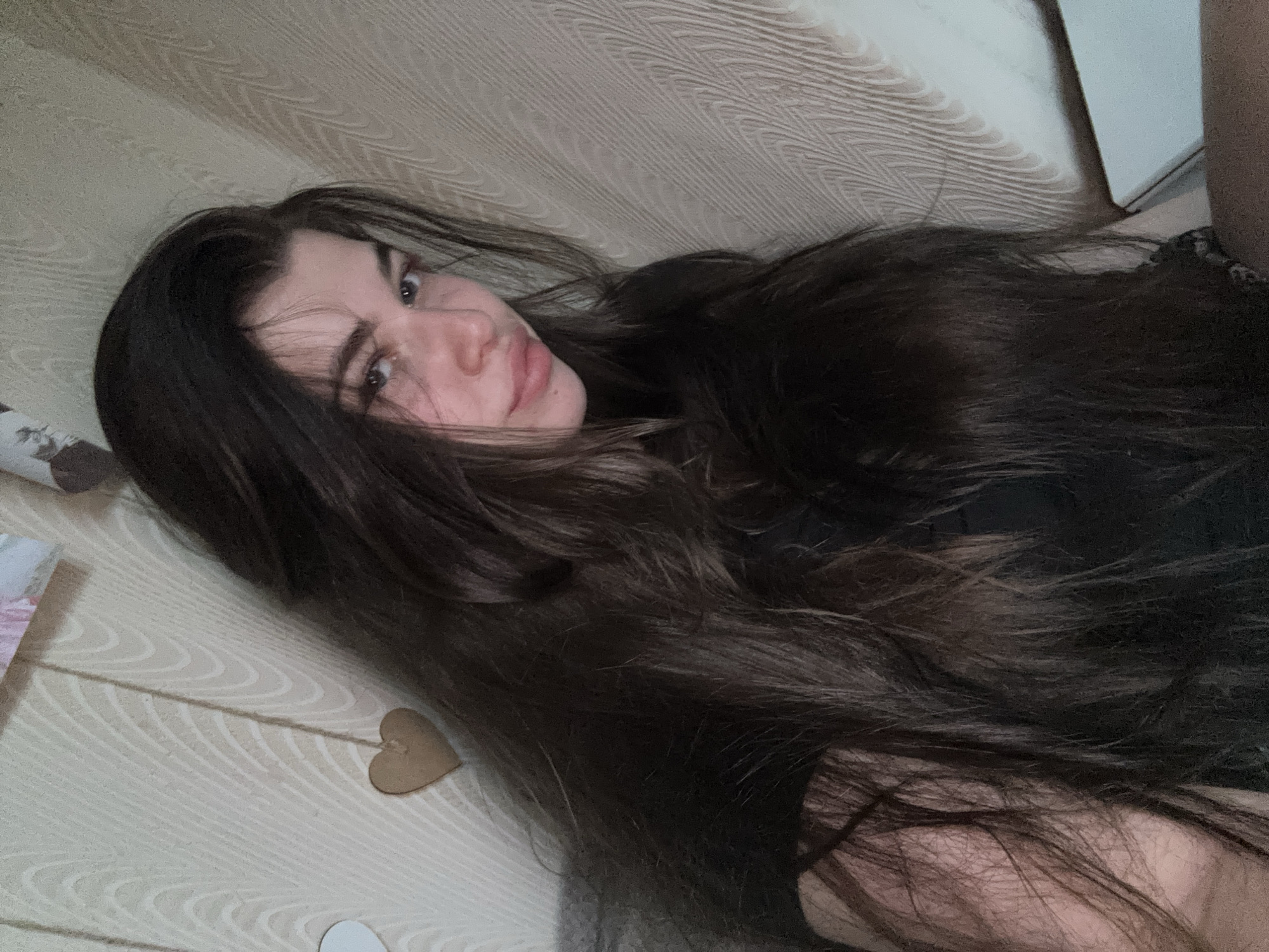 A young woman with long, dark hair is lying down on a surface with a light, patterned covering. She is looking directly at the camera with a slight smirk. A small wooden heart-shaped decoration and some paper items are visible in the lower left corner. The background is blurry, but appears to be a white wall or surface. The location of Мещёры, Russia, cannot be determined from the visual information in the image.