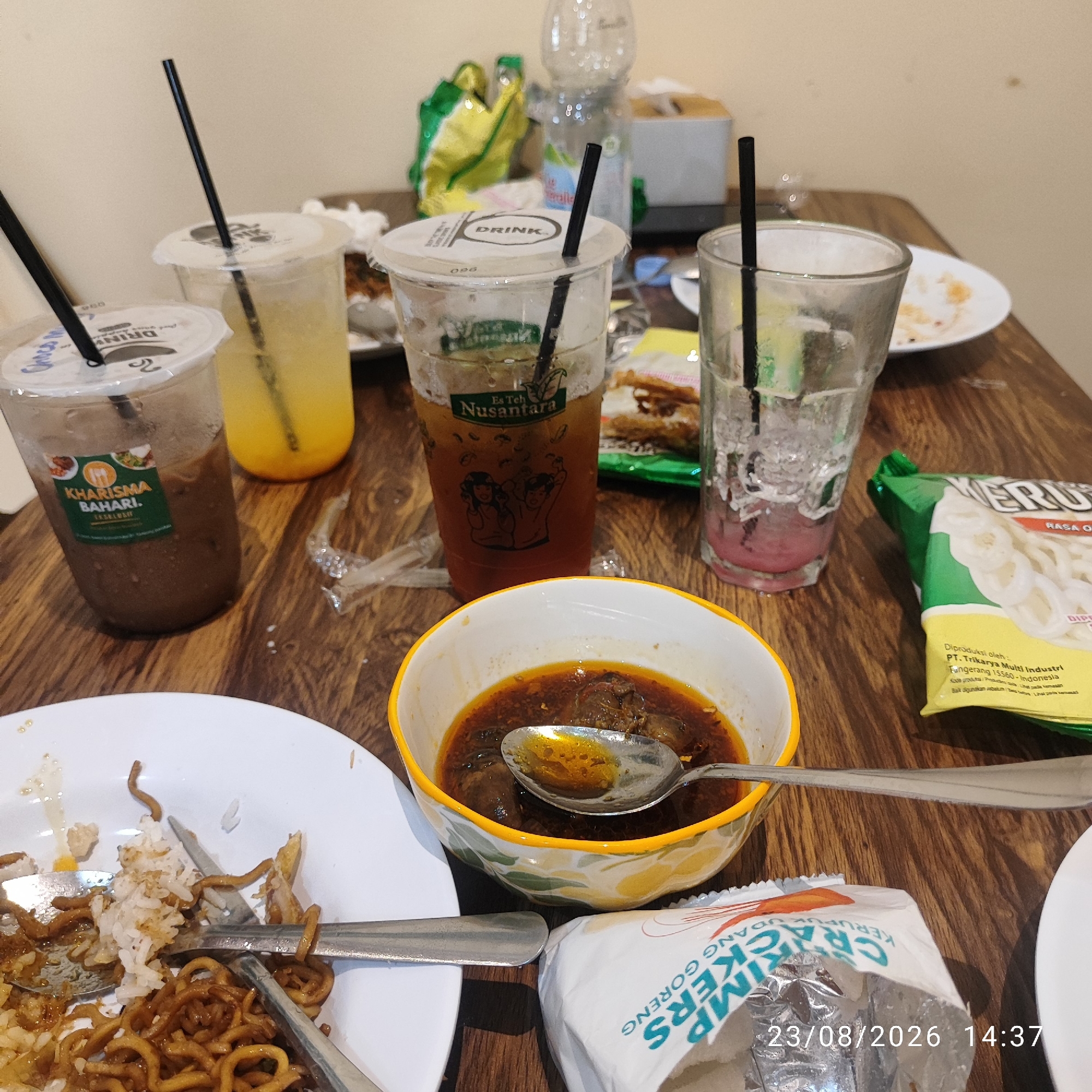 The image depicts a close-up, eye-level view of a partially eaten meal on a wooden table, likely in a casual dining setting in Tanjung Pendam, Indonesia. The scene is cluttered with various food and drink items. In the foreground, a white plate holds leftover rice, noodles, and what appears to be pieces of fried food, with a fork and spoon resting on it. To the right of the plate is a white and yellow bowl containing a dark, soupy dish with pieces of meat, and a spoon is partially submerged in it. Next to the bowl, a crumpled white and silver package of "CRACKERS UDANG GORENG" is visible, along with a partial date stamp of "23/08/2026 14:37".
Further into the image, there are several plastic cups containing beverages. One cup, to the left, is a dark chocolate drink with a "KHARISMA BAHARI" label. Another cup, to the right of that, contains a yellow liquid and has "DRINK" printed on its lid. A third clear plastic cup, further back, holds a brown liquid, possibly iced tea, with "Es Teh Nusantara" branding and an illustration of two people. To the right of this is a clear glass filled with ice and a pinkish liquid, and another empty clear glass with ice.
Behind the drinks, a green and yellow bag of what appears to be snacks is visible. In the background, out of focus, are a plastic water bottle and a white tissue box. The overall impression is of a meal that has been consumed, with remnants left on the table. The lighting is somewhat dim, suggesting an indoor environment, possibly late in the day.