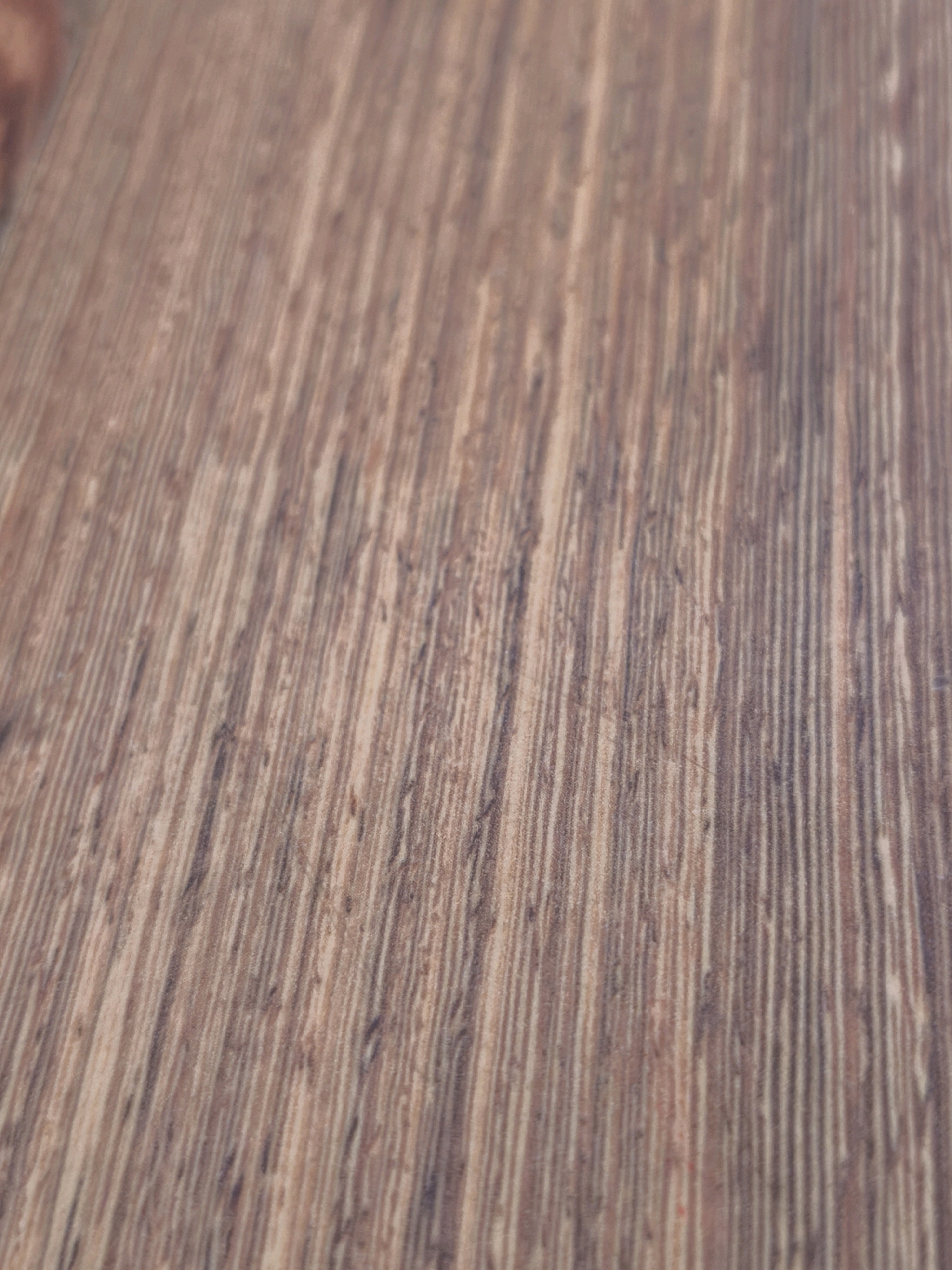 The image shows a close-up, angled view of a wooden surface, likely a floor or tabletop, with a prominent wood grain pattern. The wood is a medium brown color with darker striations running vertically and diagonally across the frame. The texture appears to be slightly rough, suggesting it might be unfinished or aged wood. The lighting is diffused, casting soft shadows and highlighting the natural variations in the wood's texture. There are no people, animals, or discernible objects present in the image. The scene is devoid of any urban or natural environmental context; it is solely focused on the material of the wood surface. This image does not contain any information that could be used to determine its location in Karachayevsk, Russia, or any other geographical area.