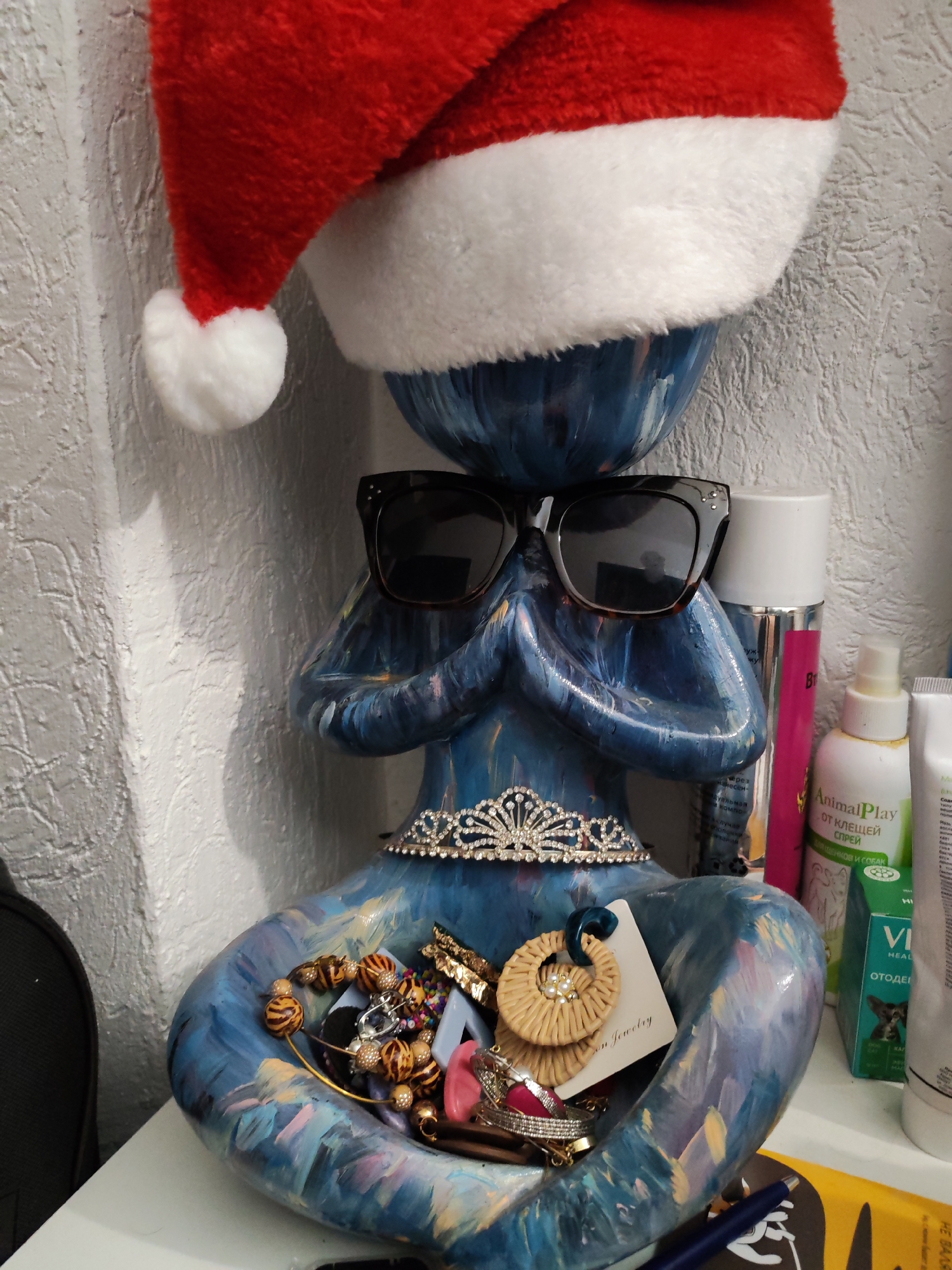A blue decorative sculpture, possibly a woman in a yoga pose, is wearing a red and white Santa Claus hat and black sunglasses. A silver tiara is placed around the sculpture’s neck. The sculpture is holding a collection of jewelry, including beaded necklaces, a gold bracelet, and earrings with a tag that says "On Jewelry." The sculpture sits on a white surface, likely a shelf or table. Behind the sculpture are various bottles and containers, including one labeled "AnimalPlay от клещей спрей" and another labeled "VI HEALTH ОТОДЕИ." A black object is visible on the left side of the image. A blue pen is visible in the bottom of the sculpture's hands. The background is a textured white wall.