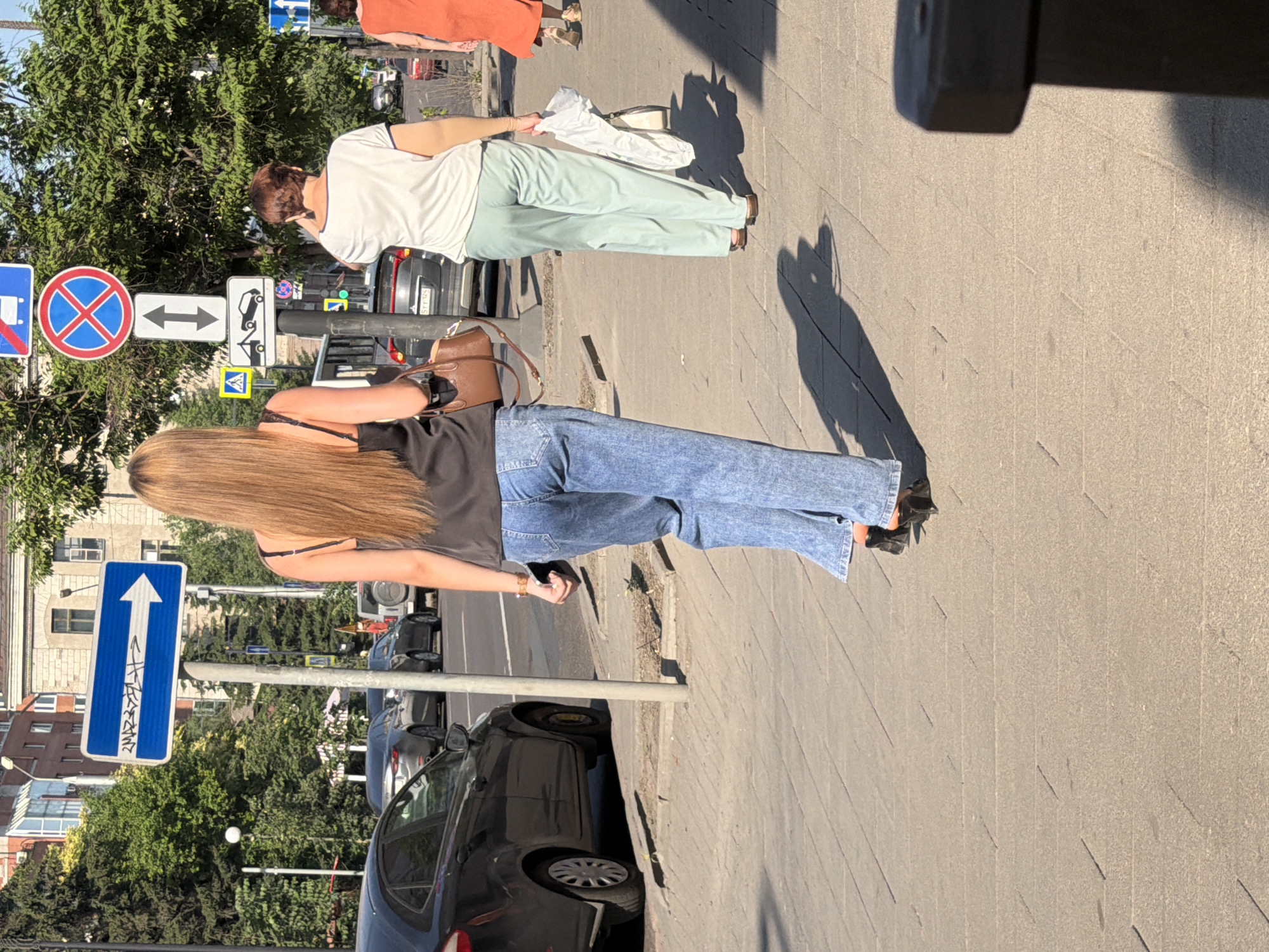 A street scene in Krasnoyarsk, Russia, features three individuals walking on a sidewalk. In the foreground, a person with long blonde hair wears a black tank top and loose-fitting blue jeans. They carry a brown handbag. To their right, a person with short brown hair and a white t-shirt walks in light green pants, holding a white bag. Further to the right, a person in an orange dress is partially visible. The background shows a street with cars and trees, along with various traffic signs, including a blue sign with a white arrow and a circular sign with a red border and a diagonal red line through it. The pavement is made of rectangular blocks, and shadows are cast across it by the people and objects in the scene, indicating a sunny day.