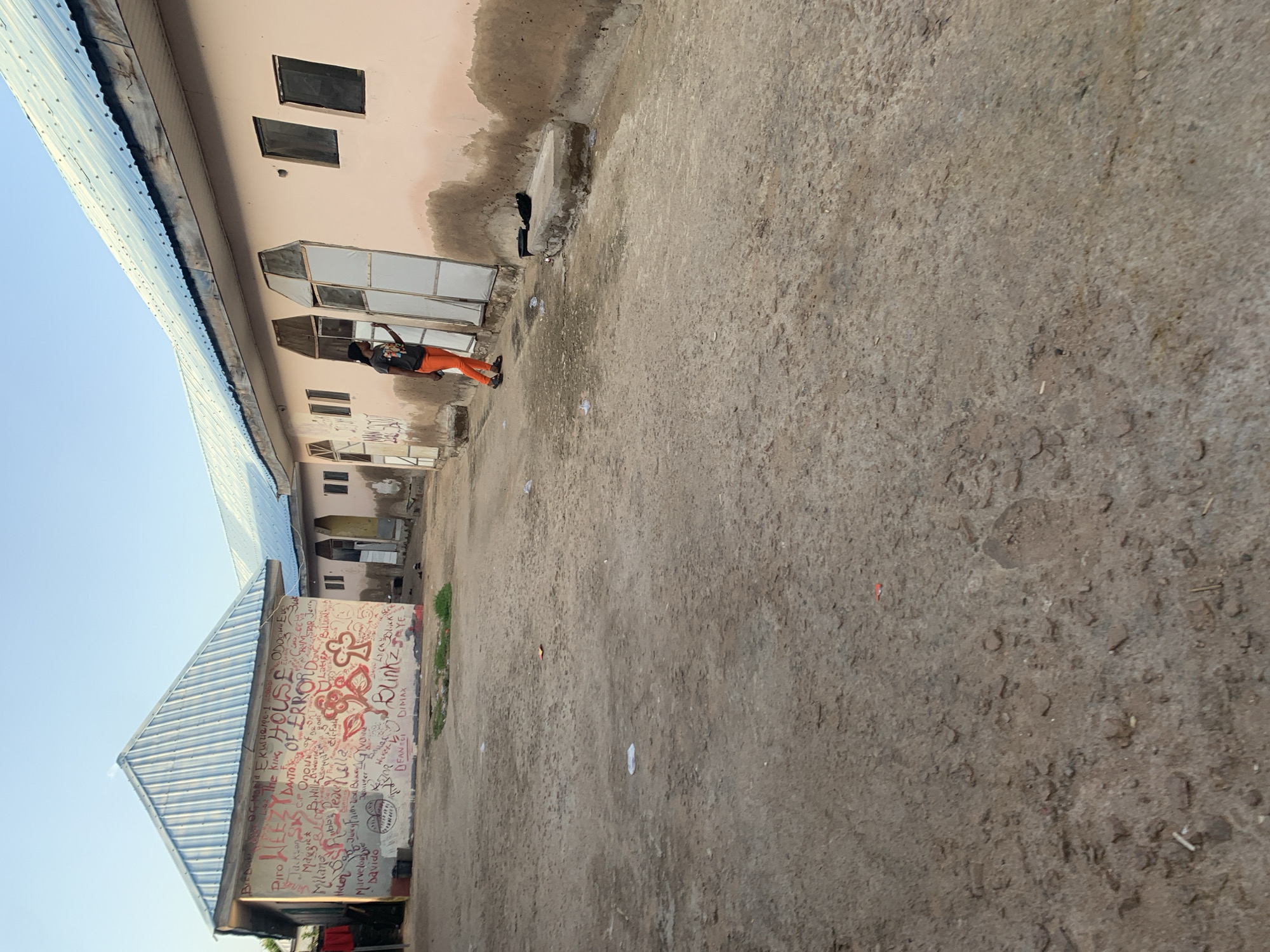 The image depicts a street scene in Jalingo, Nigeria, with a focus on a pink-colored building covered in graffiti. The building has a corrugated metal roof and several windows, some of which are covered with white material. In front of the building, the ground is unpaved and appears to be dirt. A person wearing an orange jumpsuit and a dark t-shirt is standing near the building, seemingly interacting with the wall. Trash and debris are scattered on the ground. The sky is visible in the background, suggesting it is daytime.