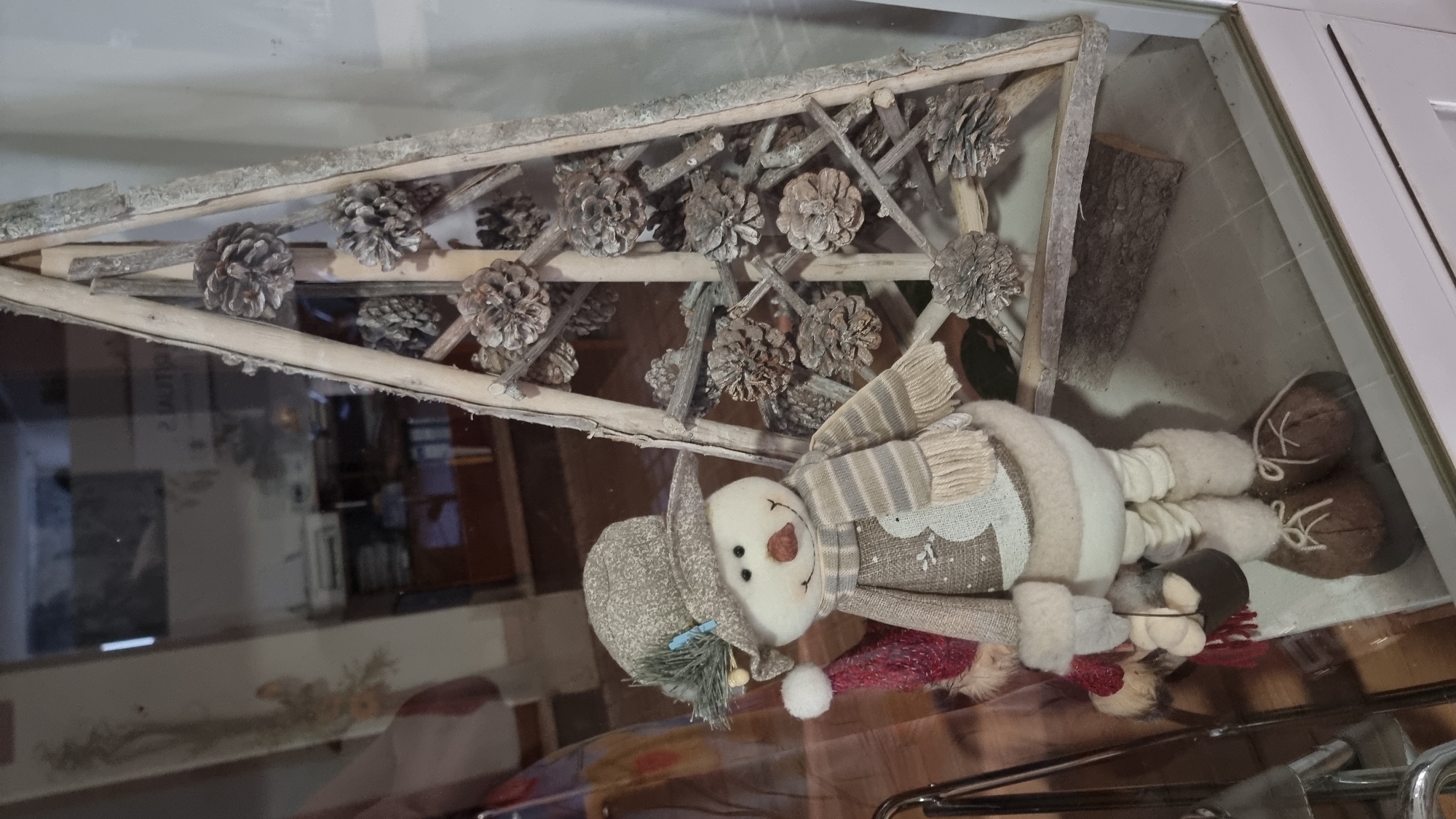 A snowman decoration stands in front of a rustic, triangular wooden structure adorned with pinecones. The snowman wears a hat, scarf, and a festive outfit. The scene appears to be a display, possibly in a shop window or home, in the city of Sant Joan Les Fonts, Spain. The background is blurred, showing reflections of an interior environment with furniture and shelves.